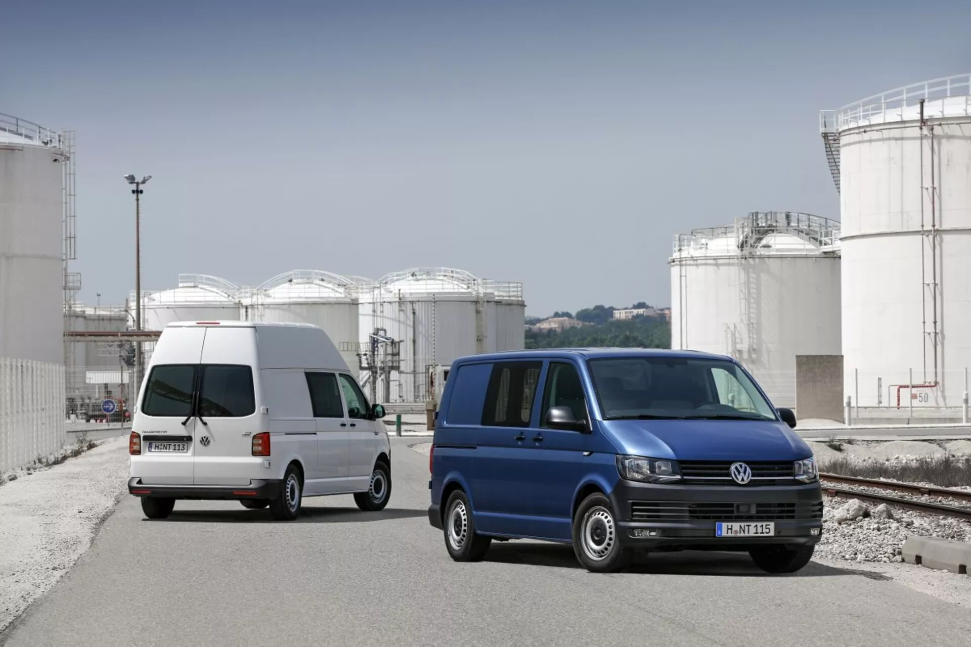 Due furgoni Volkswagen Transporter parcheggiati in un sito industriale con grandi serbatoi di stoccaggio sullo sfondo, mostrando il loro design commerciale utilitario, porte laterali scorrevoli e ruote in lega standard.