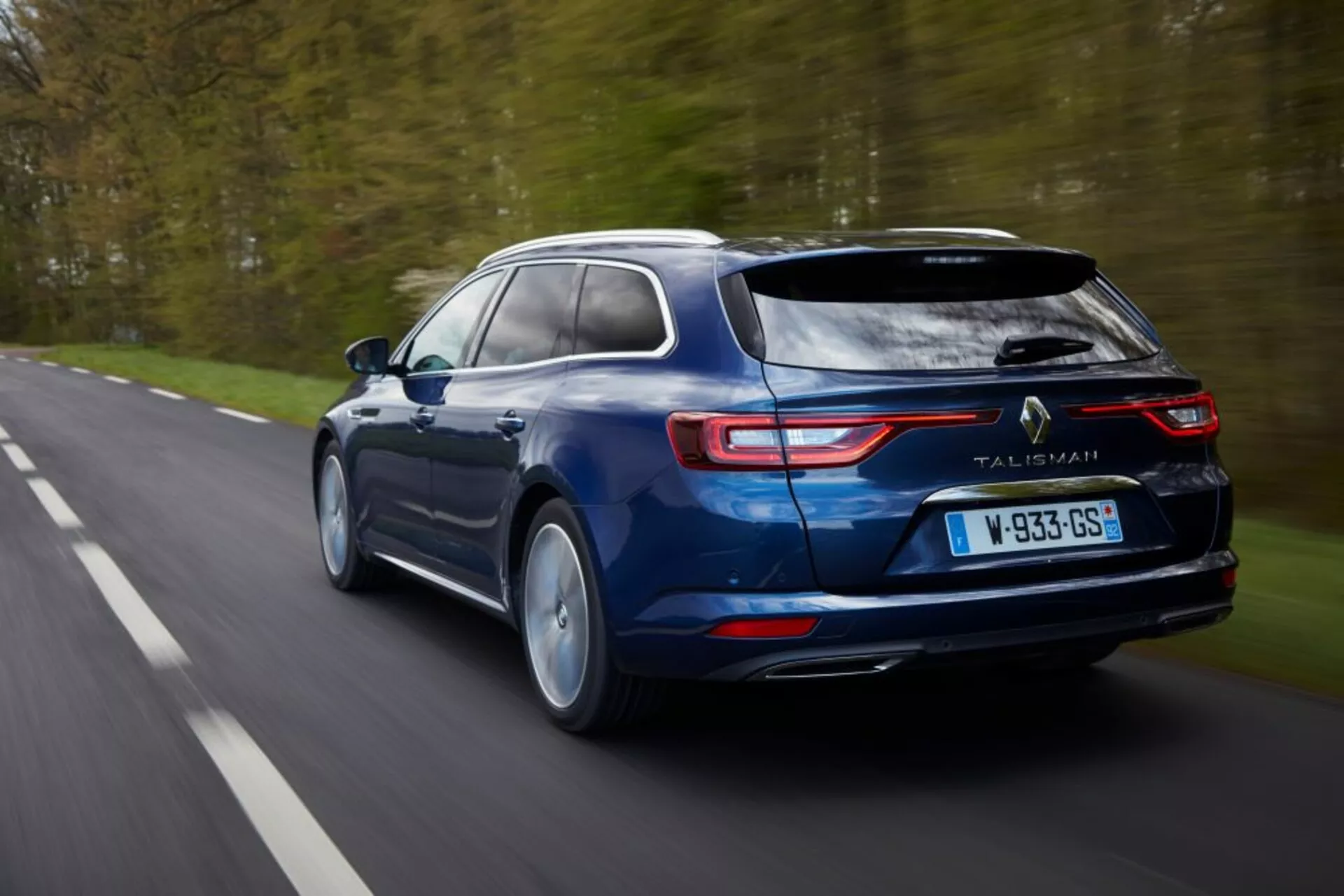 Station wagon blu Renault Talisman Grandtour 2016 in movimento, che mette in mostra caratteristiche di design come luci posteriori a LED, tubi di scappamento e la sua silhouette elegante da una prospettiva posteriore e leggermente rialzata su una strada alberata.