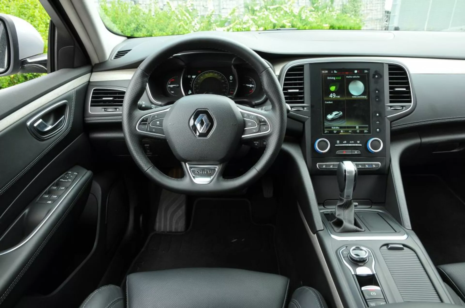 Interni della Renault Talisman 2016 che mostrano la cabina di guida con volante multifunzione in pelle, cruscotto digitale, sistema di infotainment touchscreen verticale e comandi clima eleganti dalla prospettiva del passeggero posteriore