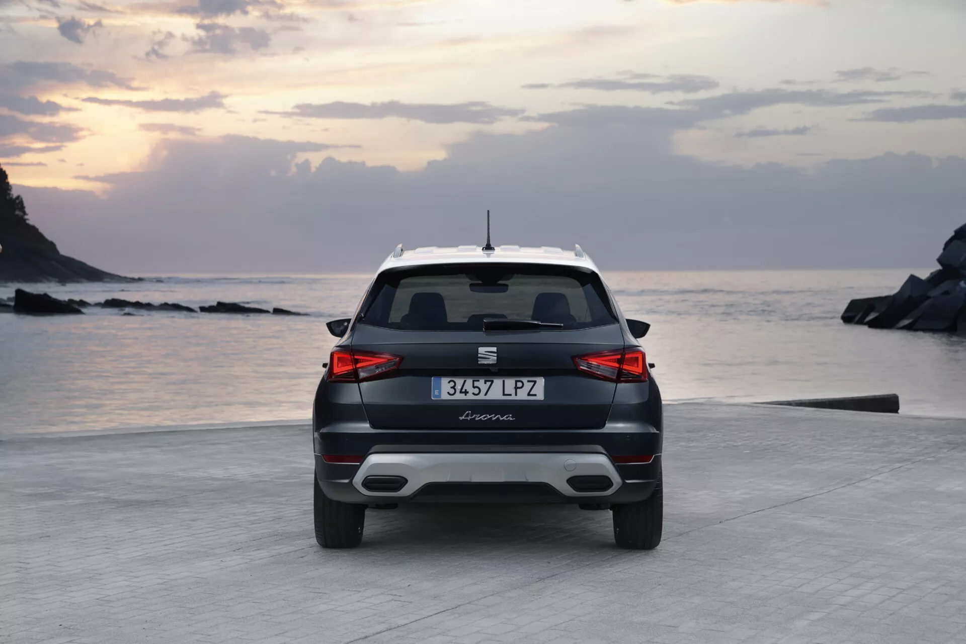 Veduta posteriore di una moderna Seat Arona 2017 all'alba o al tramonto con illuminazione soffusa, caratterizzata da spoiler posteriore sportivo, fanali posteriori orizzontali eleganti e possibili sensori di parcheggio su una strada costiera vicino alla spiaggia e alla costa rocciosa.
