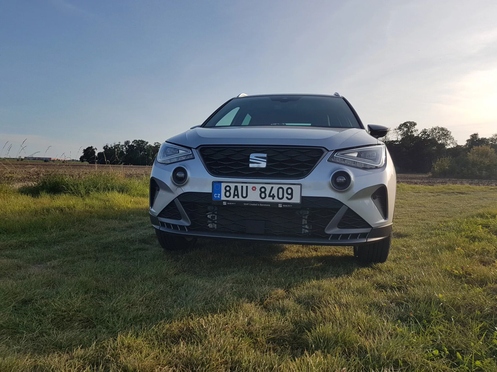 Seat Arona 2017 fotografata da un angolo frontale a livello del suolo, mostrando i suoi fari LED, griglia anteriore prominente con badge, grande presa d'aria, sensori di parcheggio sul paraurti e luci di posizione in contenitori angolari su uno sfondo di campo erboso illuminato dal tramonto