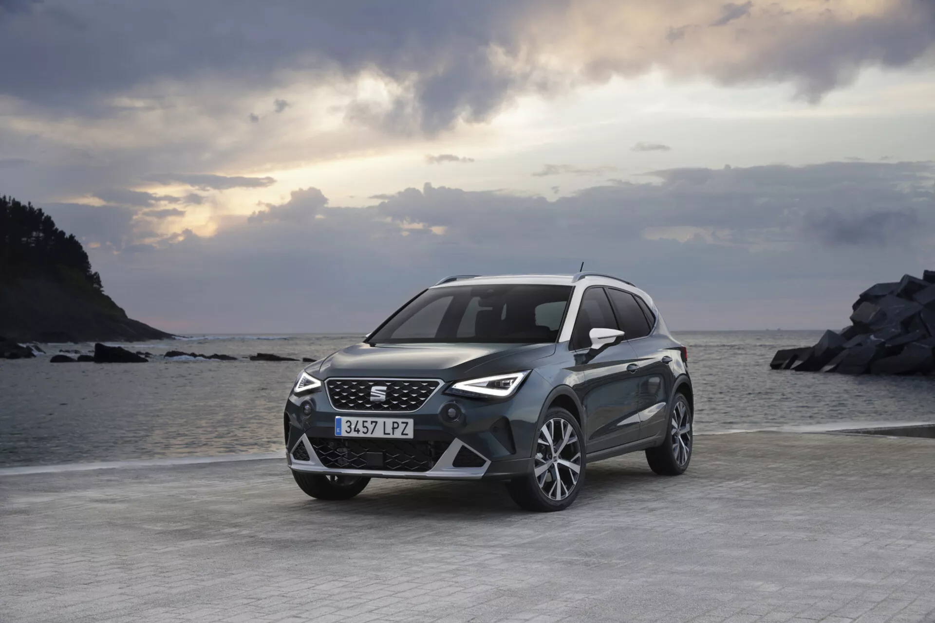 Crossover/SUV Seat Arona del 2017 parcheggiata su terreno pianeggiante con paesaggio marino e roccioso sullo sfondo durante l'alba o il tramonto con cielo parzialmente nuvoloso, evidenziando la sua vista frontale e laterale che mette in luce il design moderno della griglia e dei fari LED, paraurti anteriore sportivo, ruote in lega e finitura metallica.