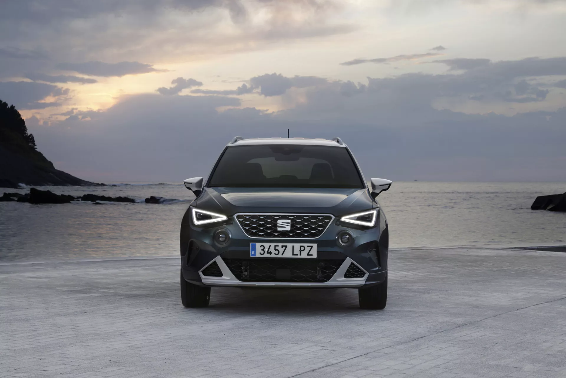 Seat Arona 2017 mostrata da un angolo frontale che mette in evidenza la sua moderna griglia con motivo a nido d'ape, fari LED, paraurti inferiore dettagliato e cerchi in lega, parcheggiata su una superficie pavimentata vicino a una spiaggia con l'oceano e il cielo crepuscolare sullo sfondo.