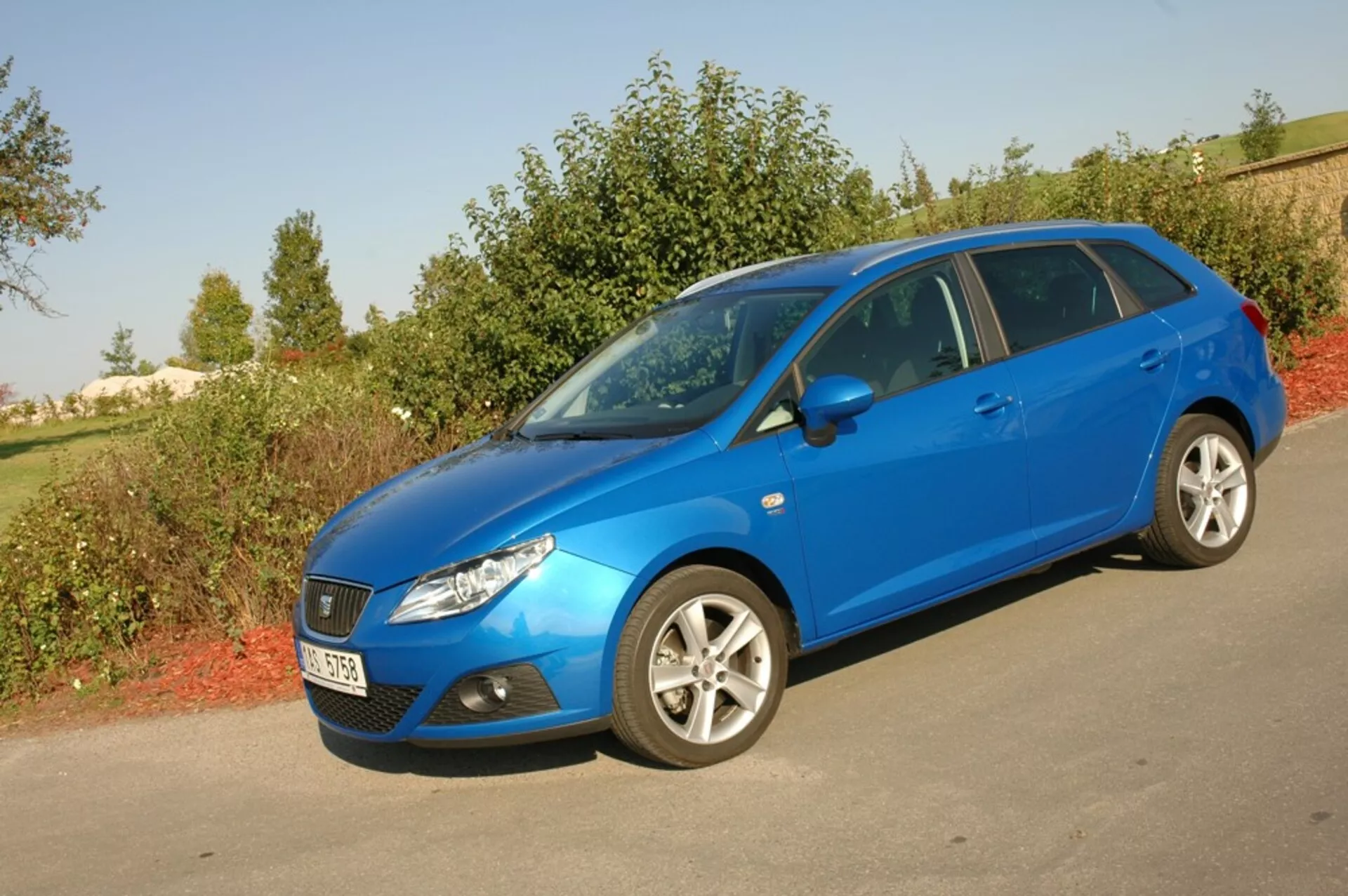 Seat Ibiza 2012 hatchback blu acceso parcheggiata su asfalto contro la vegetazione, mettendo in mostra la sua griglia anteriore aerodinamica, fari inclinati all’indietro e profilo laterale con cerchi in lega a più razze sotto la luce naturale del giorno