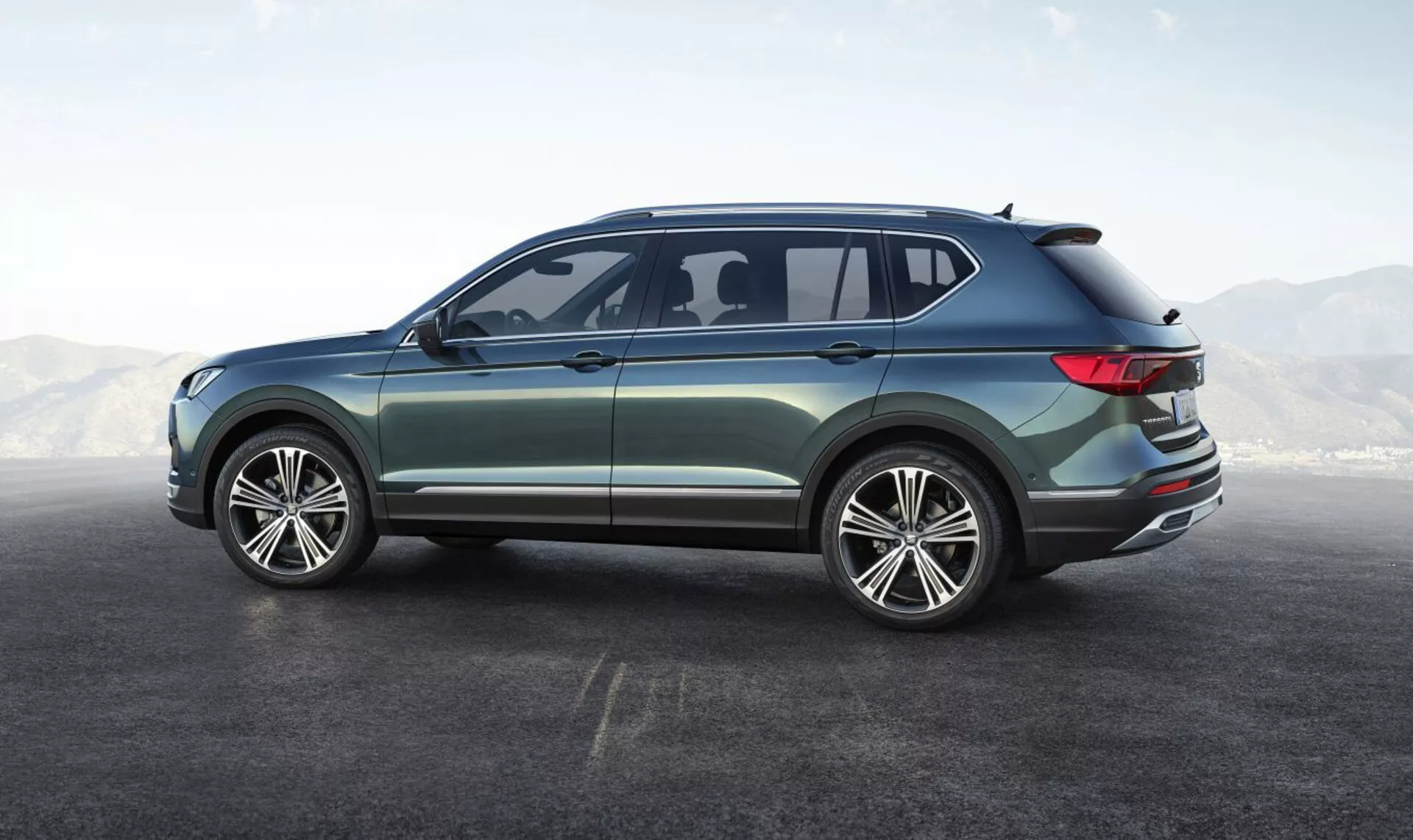SUV Seat Tarraco 2019 mostra il suo angolo posteriore laterale, caratterizzato da grandi ruote in lega a più razze, design a quattro porte, eleganti finestrini laterali con una piccola finestra posteriore aerodinamica, linea del tetto inclinata dolcemente con spoiler integrato, fanali posteriori LED avvolgenti, vernice metallica con rifiniture esterne cromate, evidenziando il suo design moderno e versatile