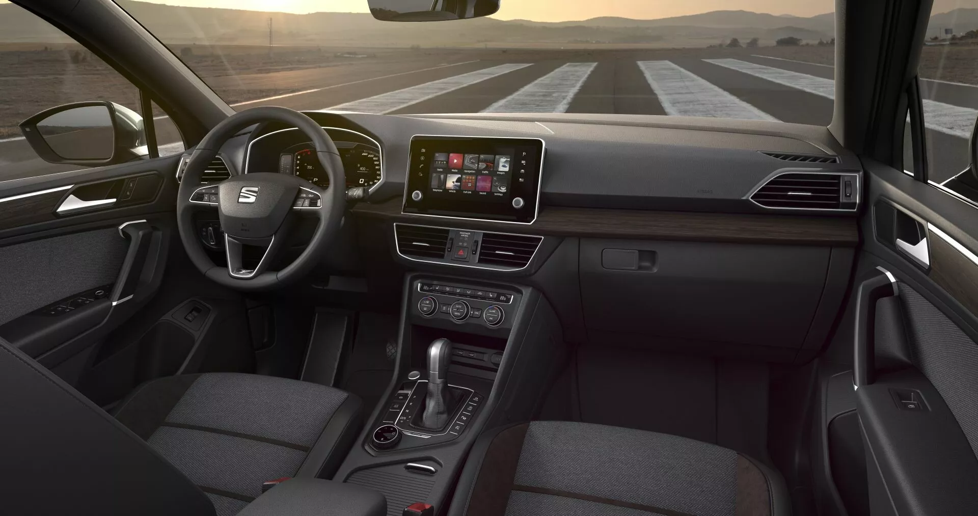 Prospettiva interna del Seat Tarraco 2019 dalla seduta posteriore, evidenziando il volante multifunzione, il display touchscreen centrale per l'infotainment, i pulsanti per il controllo del clima, la leva del cambio automatico e il sedile del passeggero in tessuto, sotto una luce interna tenue