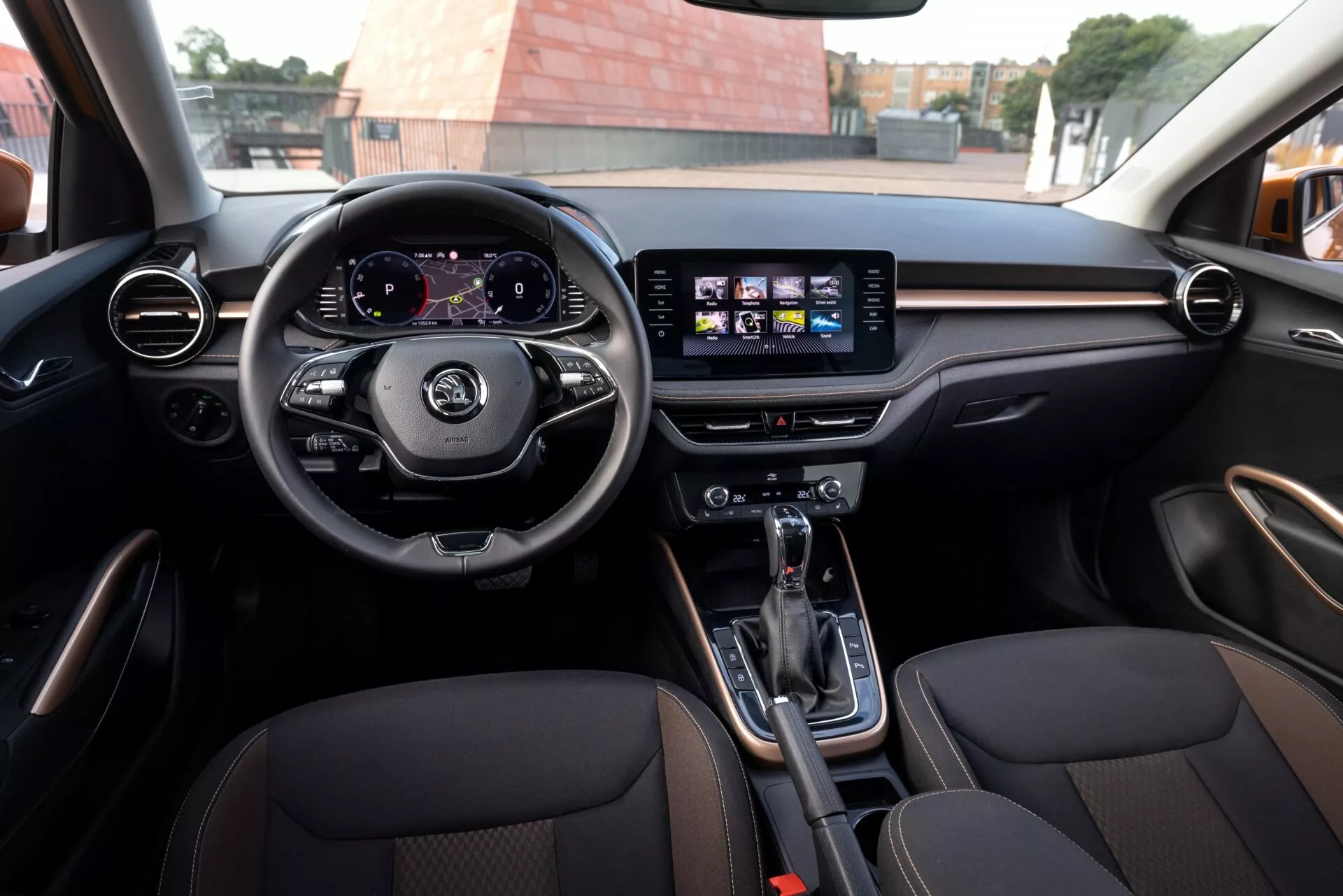 Vista dell'interno della Skoda Fabia 2021 dal sedile posteriore, che mostra un volante a tre razze con comandi, quadro strumenti digitale che visualizza tachimetro e contagiri e sistema di infotainment centrale con touchscreen con icone di navigazione e musica. Prese d'aria circolari e orizzontali con finitura che imita legno o metallo, cambio automatico con freno di stazionamento elettronico nella console centrale, sedili anteriori sportivi rivestiti in pelle con cuciture a contrasto. Ambiente moderno e spazio interno ampio indicano lusso e sportività.