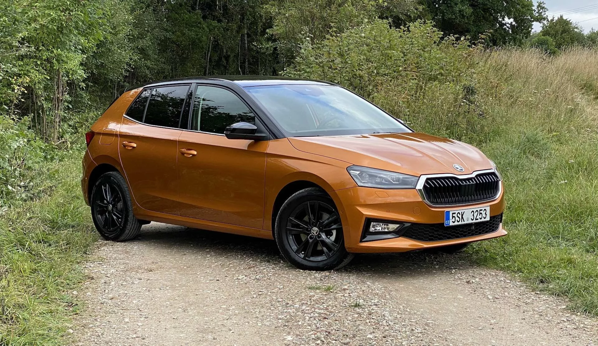 Immagine di una moderna Škoda Fabia 2021 hatchback, catturata da una prospettiva anteriore a tre quarti, che mostra il lato anteriore sinistro, la porta del conducente, il cofano sinistro e una parte del tetto. Il veicolo si trova su un sentiero sterrato circondato da vegetazione, indicando un'ambientazione rurale. Le caratteristiche visibili includono una griglia orizzontale con emblema centrale, fari LED affilati, vernice arancione metallizzata, specchietti laterali in tinta con la carrozzeria, cerchi in lega a razze multiple nere con pneumatici scolpiti e porte sagomate con linee aerodinamiche. L'auto sembra nuova, compatta e non modificata.