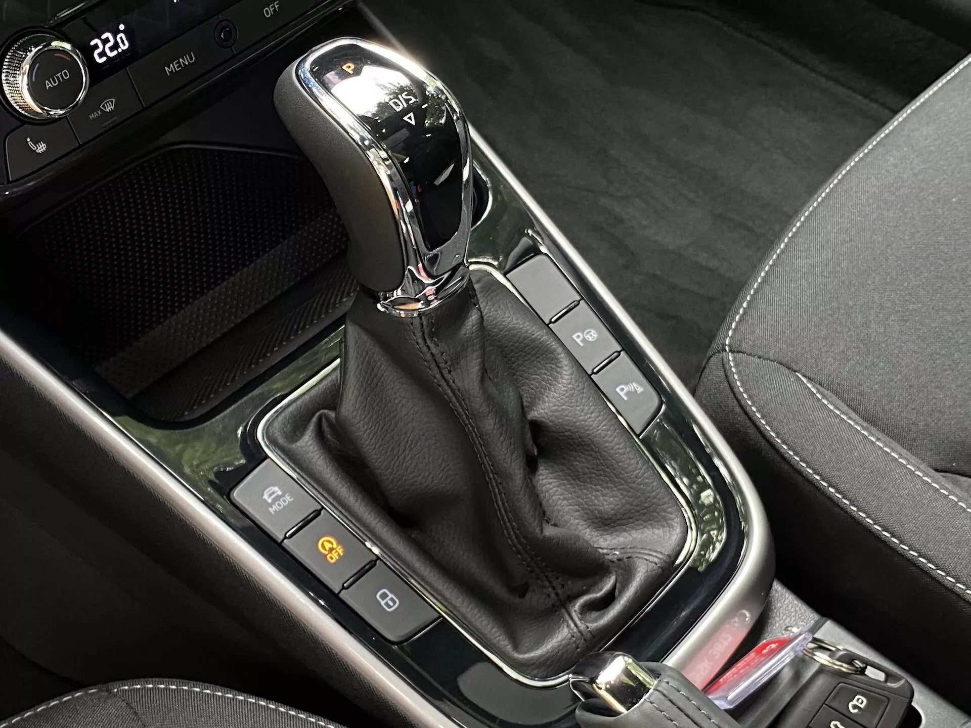 Vista ravvicinata della leva del cambio automatico nella console centrale di una Skoda Fabia 2021. La leva del cambio moderna, posizionata verticalmente in modalità 'P' (Parcheggio), presenta una finitura cromata o metallica nella parte superiore ed è incassata in un pannello nero lucido. In basso, un manicotto di pelle nera nasconde il meccanismo. Intorno alla leva del cambio c'è una serie di pulsanti e indicatori, inclusi due a sinistra probabilmente relativi alle caratteristiche di sicurezza dell'auto, e parte di un pulsante rosso a destra. L'area mescola vari materiali neri, comprese superfici lucide e modelli che imitano la fibra di carbonio, suggerendo un design contemporaneo ad alta tecnologia.