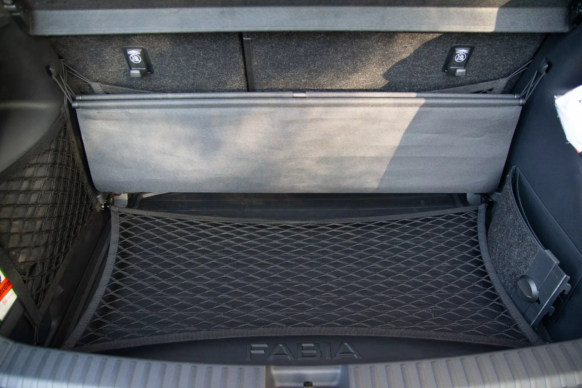 Vista dall'alto dell'area del bagagliaio di una Skoda Fabia 2021, mostrando il compartimento di stivaggio interno dettagliato con una rete da carico sul lato sinistro per il fissaggio degli oggetti e un pannello del pavimento flessibile parzialmente sollevato che rivela uno spazio di stivaggio aggiuntivo sotto. Il bagagliaio presenta una moquette durevole e una piastra di soglia con il marchio 'FABIA' per la protezione durante il carico e lo scarico.