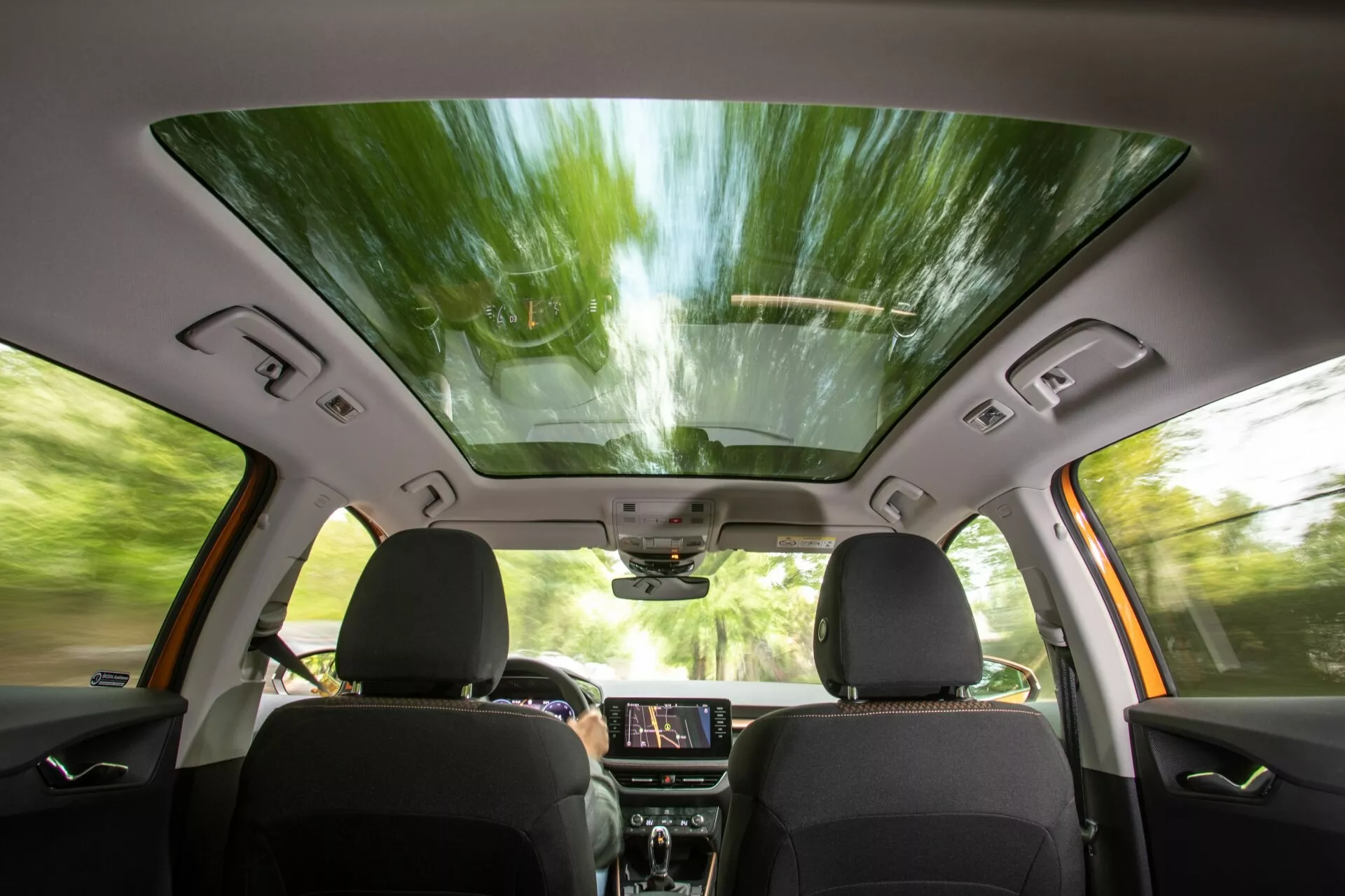 Vista interna della Skoda Fabia 2021 dalla prospettiva del sedile posteriore, che mostra i sedili anteriori con rivestimento scuro, tetto panoramico, schermo touchscreen per l'infotainment illuminato sul cruscotto, pannelli di controllo, altoparlanti integrati nelle porte e comandi dei finestrini, specchietto retrovisore con possibili funzioni high-tech e una console superiore per controlli aggiuntivi, suggerendo un'esperienza di guida moderna, confortevole e migliorata dalla tecnologia.