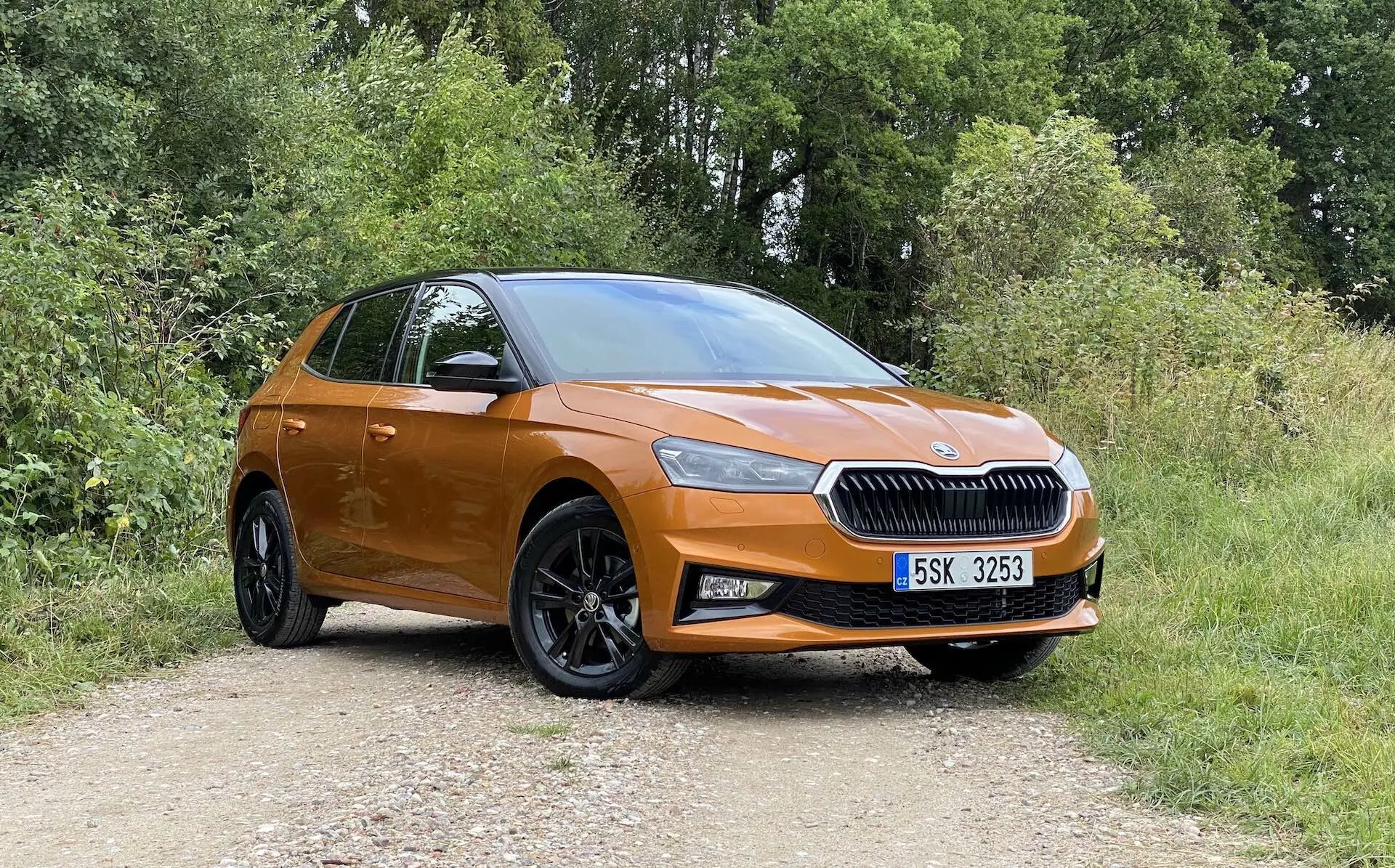 Profilo laterale completo di una moderna hatchback Skoda Fabia 2021 di colore arancione parcheggiata su un sentiero sterrato con vegetazione sullo sfondo, catturata da un angolo leggermente obliquo dal davanti, evidenziando il lato del conducente. Caratteristiche includono una griglia prominente con il logo del marchio centrato, fari eleganti, luci fendinebbia, grandi cerchi in lega nero a più razze, maniglie e specchietti in tinta con la carrozzeria, linea del tetto inclinata e un accenno al portellone posteriore e al gruppo luci.
