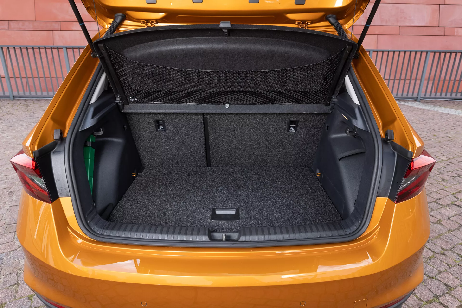 L'immagine mostra il bagagliaio di una Skoda Fabia modello 2021, ripreso da un angolo posteriore leggermente elevato per mostrare lo spazioso vano di carico. L'interno è rivestito con un materiale simile a un tappeto e presenta aree di stoccaggio laterali flessibili, una rete di divisione per il carico e un'area incassata nella parte inferiore per ulteriore spazio di stivaggio. Il portellone è sollevato, rivelando un grande bagagliaio pulito, progettato per sottolineare la capacità di carico e la praticità del veicolo.