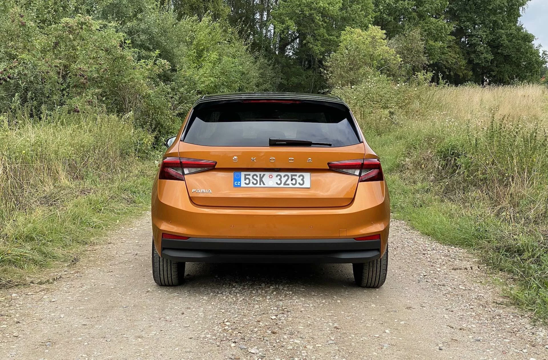 Vista posteriore di una Skoda Fabia 2021 hatchback arancione metallizzato brillante fotografata alla luce del giorno su un sentiero stretto in un ambiente rurale. L'immagine cattura il design posteriore moderno con una barra di luce posteriore continua, fanali angolari, riflettori integrati, tergicristallo posteriore e spoiler posteriore per l'aerodinamica, evidenziando la finitura lucida e le caratteristiche pratiche dell'auto.