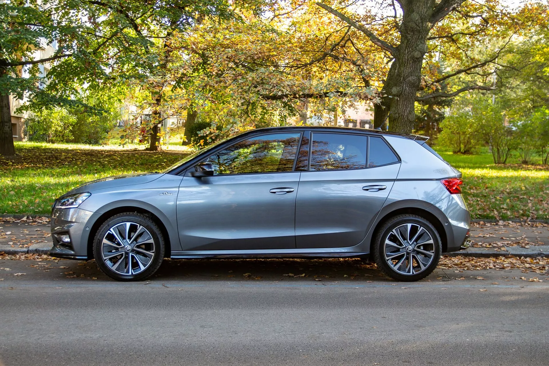 Vista laterale di una Skoda Fabia 2021 color argento parcheggiata accanto a una strada fiancheggiata da alberi durante il giorno, mostrando il suo design moderno con linea di cintura alta, cerchioni in lega sportivi, maniglie delle porte a filo, specchietti laterali aerodinamici con indicatori di direzione integrati, linea del tetto inclinata, linee di carrozzeria nitide, finestre parzialmente oscurate e montante B nero. L'ambiente sereno suburbano o del parco esalta l'estetica del veicolo.