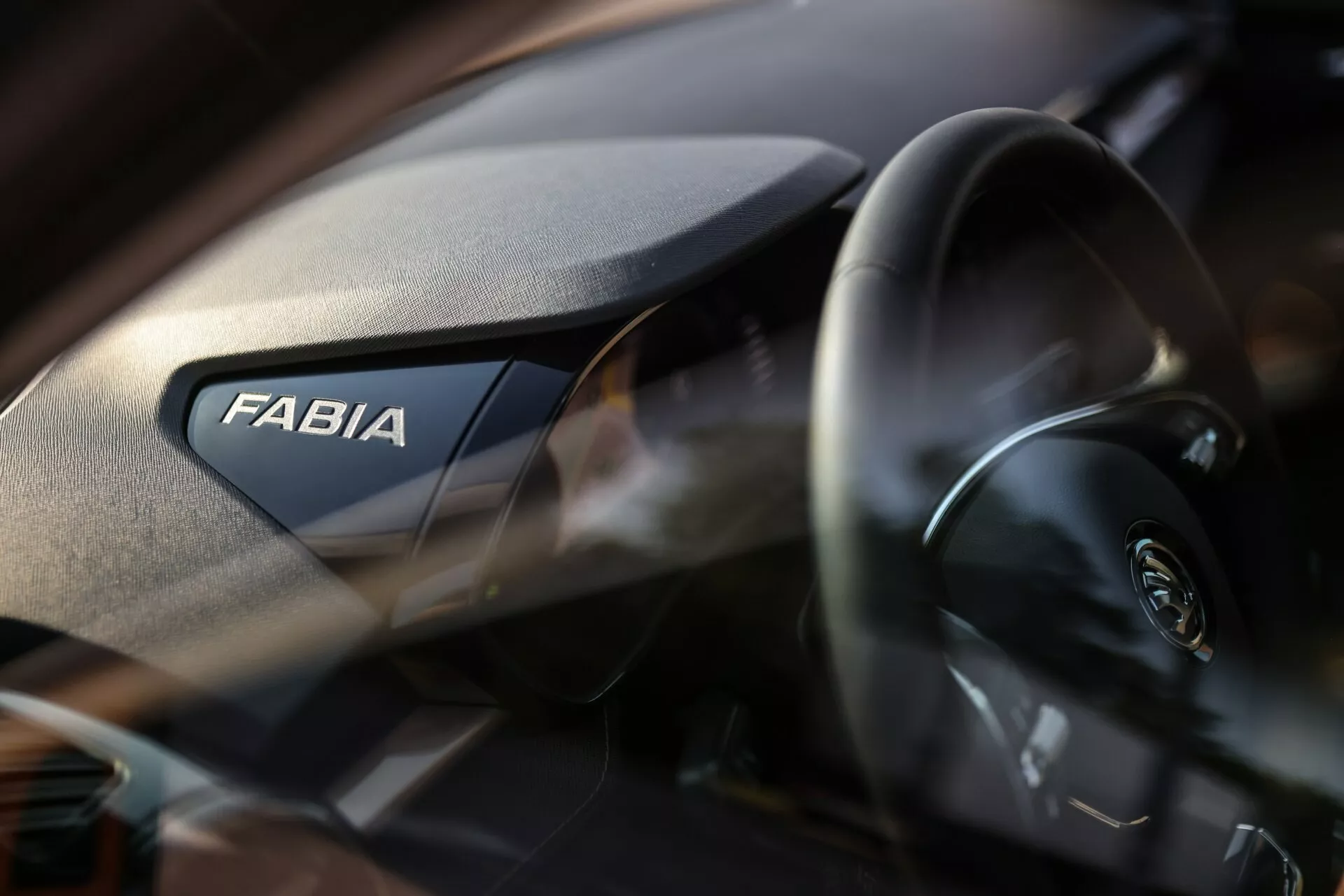 Vista ravvicinata degli interni della Skoda Fabia 2021, con evidenziato il lato passeggero del cruscotto. La parola 'FABIA' è visibile in modo prominente, mentre il volante moderno con il logo Skoda è parzialmente visibile a sinistra. Il cruscotto presenta una texture granulata e la bassa profondità di campo enfatizza il marchio con uno sfondo leggermente sfocato.
