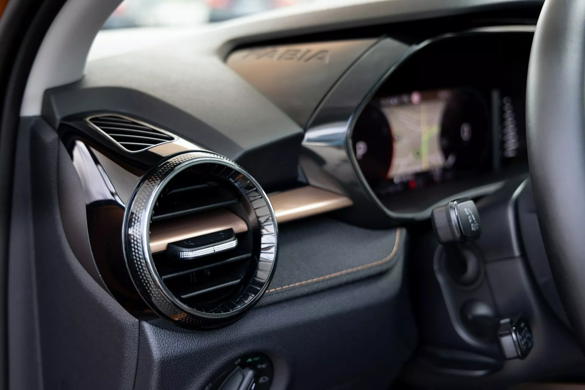 Vista ravvicinata dell'interno della Škoda Fabia 2021 focalizzata sul lato del passeggero. L'immagine cattura una bocchetta dell'aria rotonda con una finitura metallica o cromata contro un pannello testurizzato in fibra di carbonio, indicando un allestimento sportivo o di lusso. A destra è visibile il bordo di uno schermo touchscreen di infotainment integrato nel cruscotto. Sotto la bocchetta sono presenti vari pulsanti di controllo, probabilmente per l'audiosistema, il climatizzatore o altre funzioni del veicolo. La profondità di campo ridotta mette in risalto la bocchetta, suggerendo delicatamente i componenti tecnologici circostanti.