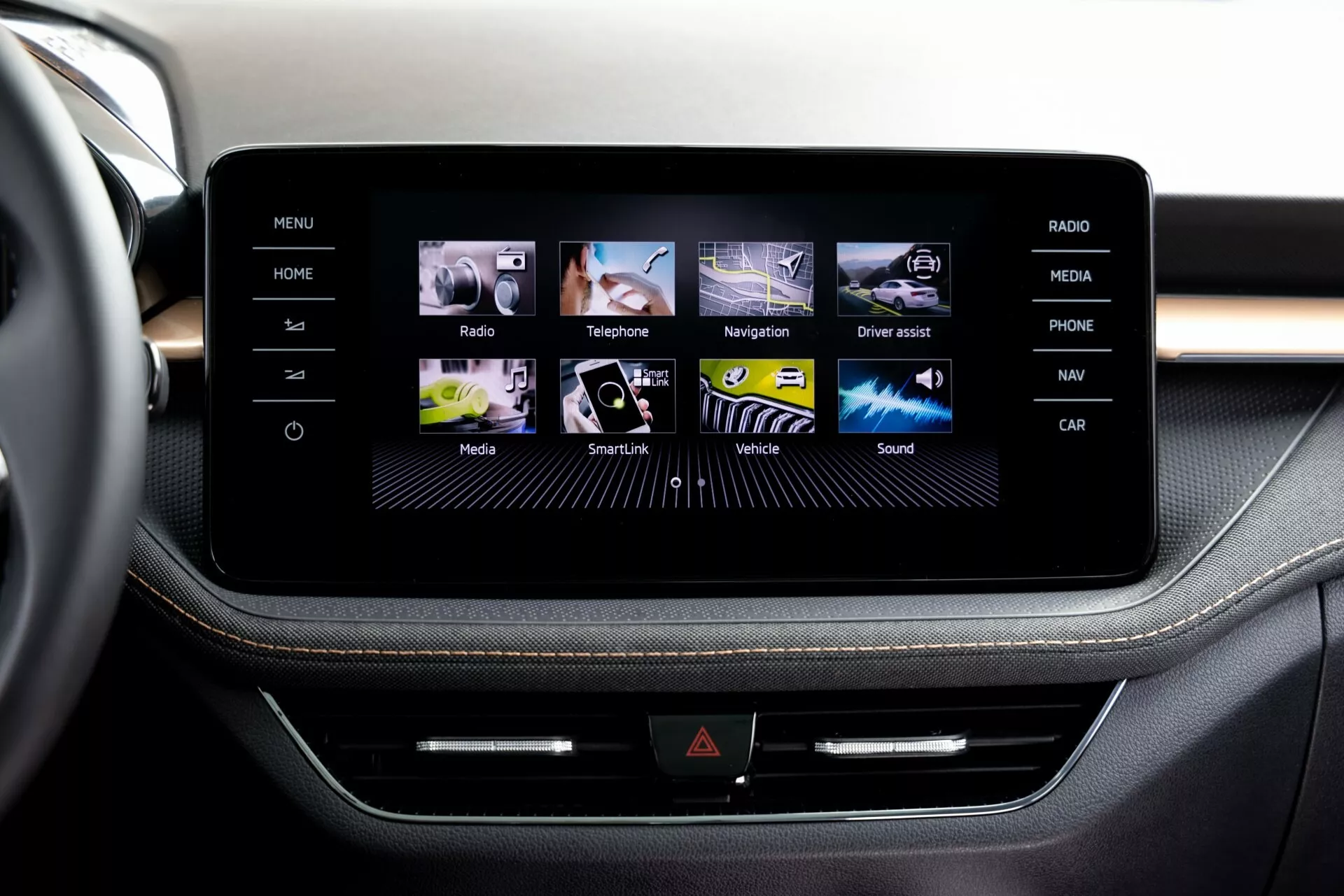 Vista ravvicinata del sistema di infotainment touchscreen della Škoda Fabia 2021 dalla prospettiva di un passeggero o conducente nella fila anteriore, che mostra una consolle centrale elegante incorniciata da quello che sembra essere alluminio spazzolato. Lo schermo visualizza un'interfaccia grafica intuitiva con icone colorate per Home, Radio, Telefono, Navigazione e altro. Sotto lo schermo, una fila di pulsanti fisici e una possibile fessura per CD aggiungono multifunzionalità. I rivestimenti premium con cuciture a contrasto suggeriscono un design degli interni di alta gamma.