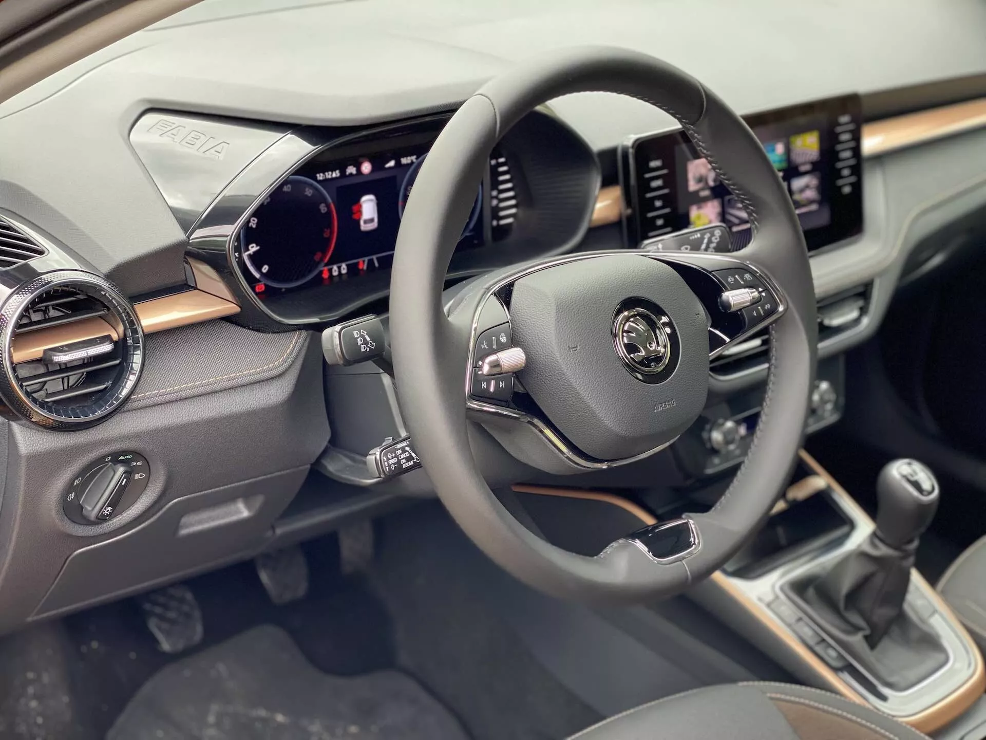 Vista interna della Skoda Fabia 2021 focalizzata sul lato del conducente, mostrando il volante centrato con comandi multifunzionali per audio, comandi vocali e controllo della velocità. Il quadro strumenti digitale visualizza tachimetro, contagiri e indicatori di stato del veicolo. Un sistema di infotainment touchscreen è visibile sul cruscotto con funzionalità di navigazione e media. Sotto lo schermo ci sono manopole e pulsanti per il controllo del clima. La leva del cambio manuale è visibile nella console centrale.