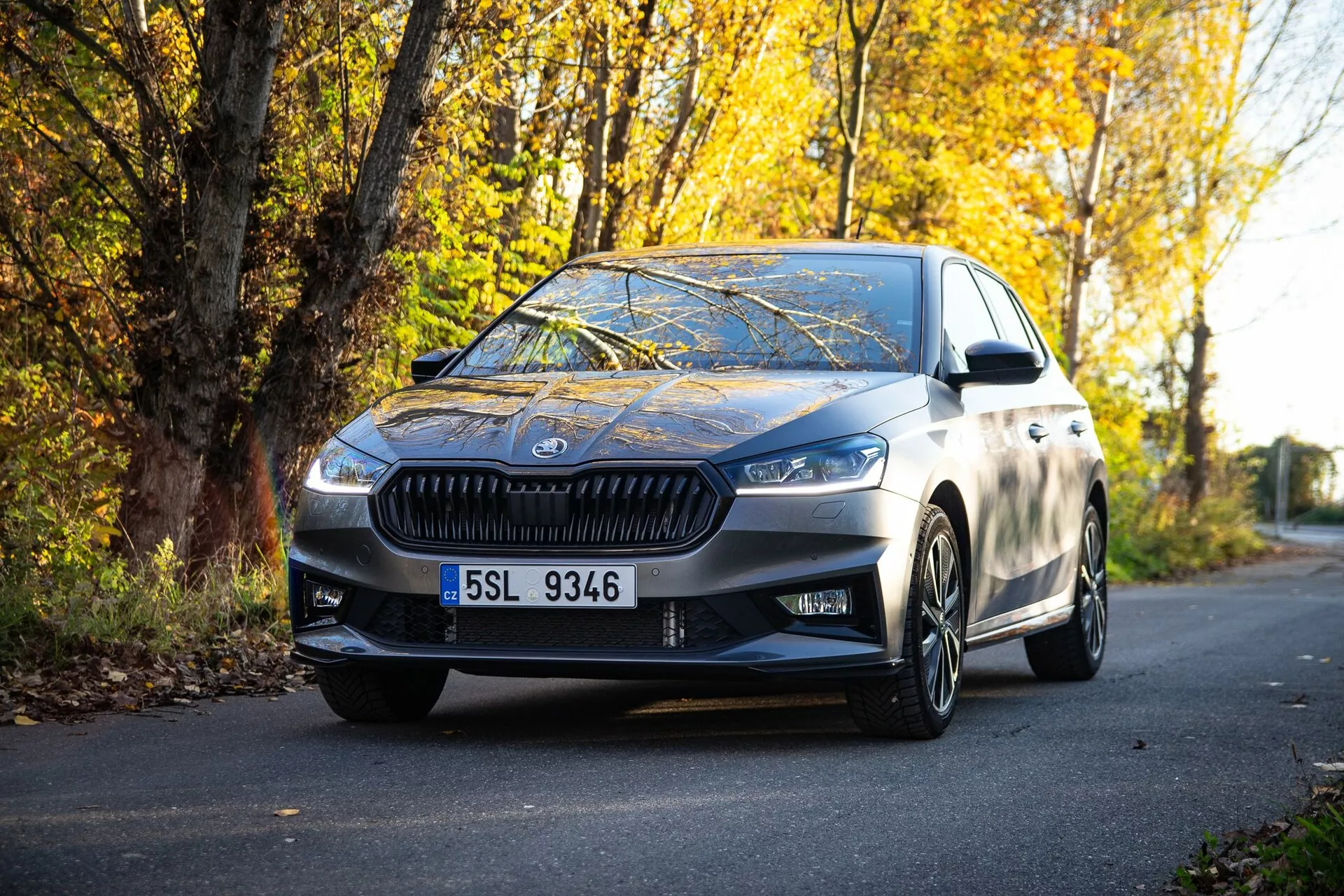 Immagine di una moderna berlina Skoda Fabia 2021 parcheggiata su una strada fiancheggiata da foglie autunnali, catturata da una prospettiva a livello del suolo, evidenziando la griglia anteriore, i fari angolari, le linee di design eleganti, i cerchi in lega e gli specchietti laterali con indicatori di direzione integrati, riflettendo una finitura metallica di alta qualità e una combinazione di lusso e sportività.