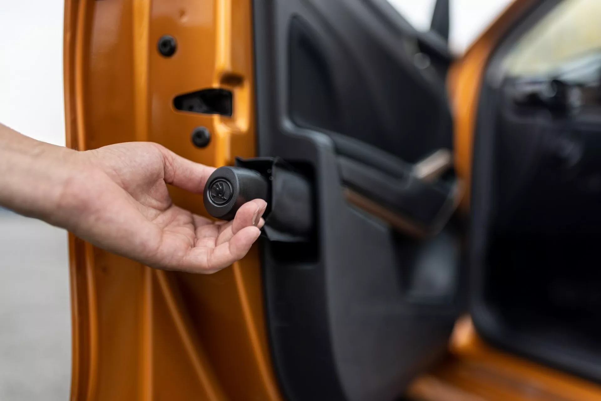 Primo piano della porta del conducente della Skoda Fabia 2021, leggermente aperta, vista dall'esterno con il bordo interno visibile. Una mano tiene un compatto portachiavi nero e circolare che indica un sistema di accesso senza chiave o di sicurezza dell'auto. L'auto è di colore arancione brillante con una finitura metallica, che mostra meccanismi di blocco avanzati e caratteristiche di sicurezza personalizzate.