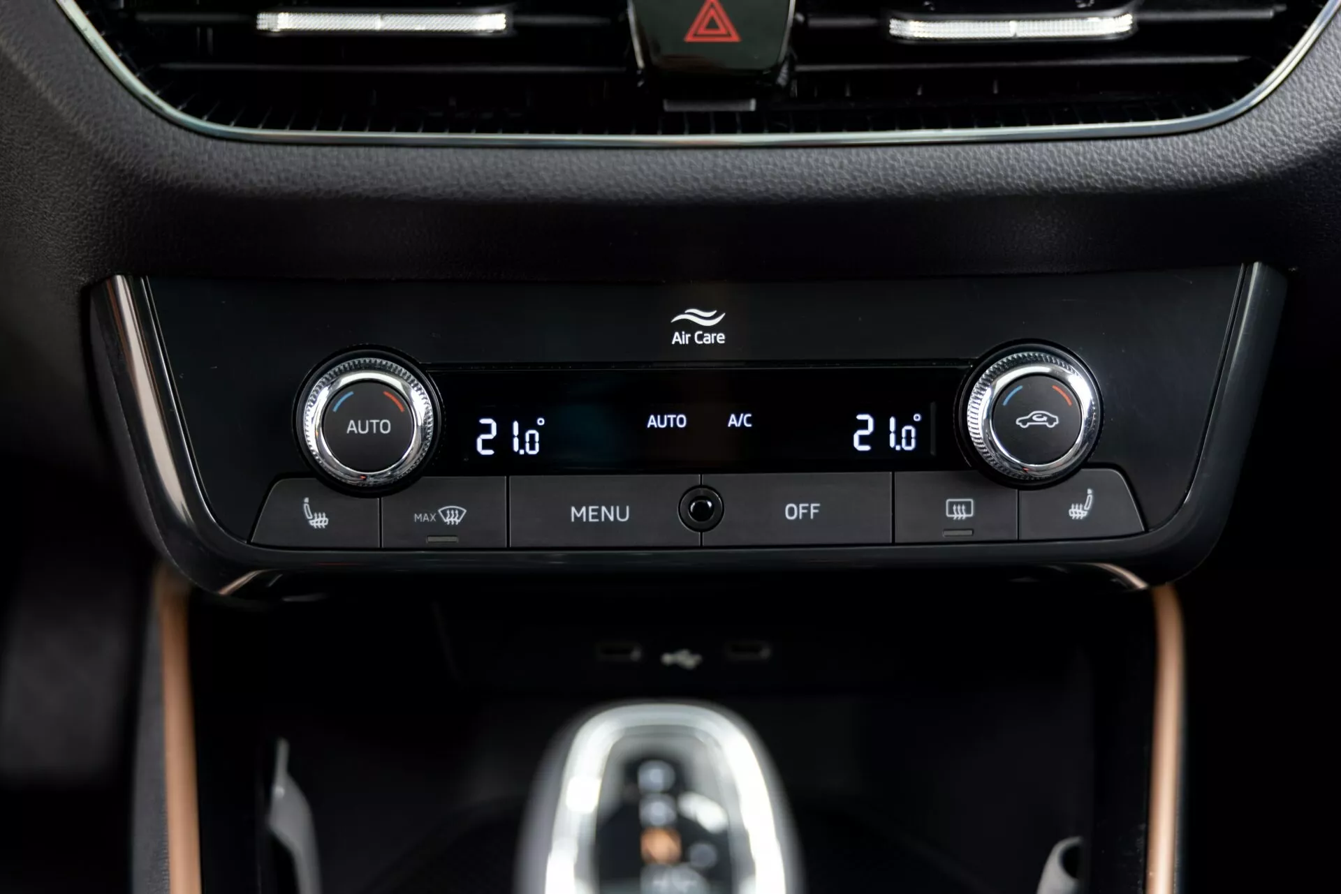 Vista ravvicinata del pannello di controllo del climatizzatore della Skoda Fabia 2021 situato sulla console centrale, con controlli della temperatura a doppia zona con display digitali che mostrano '20', pulsanti integrati per il controllo automatico del clima ('AUTO'), aria condizionata ('AC'), impostazioni del menu ('MENU') e spegnimento del sistema ('OFF'). Una cornice nera lucida circonda un display informativo sopra i controlli, con un pulsante di avvertimento di pericolo (icona triangolare) visibile in alto. Il pannello è inserito in un interno premium con finiture opache e effetto legno, evidenziando un design e una tecnologia moderni.