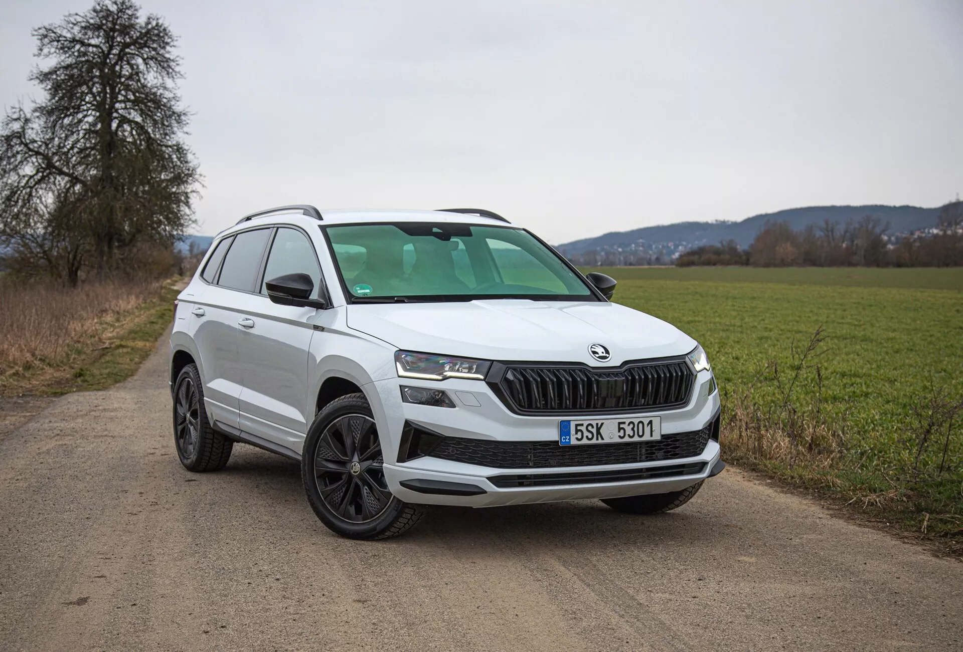 SUV bianco Škoda Karoq del 2021 parcheggiato su una strada di terra di campagna, mostrando la vista del quarto anteriore sinistro inclusi griglia, fari e design delle ruote, con focus su caratteristiche moderne come illuminazione LED e sensori di parcheggio.