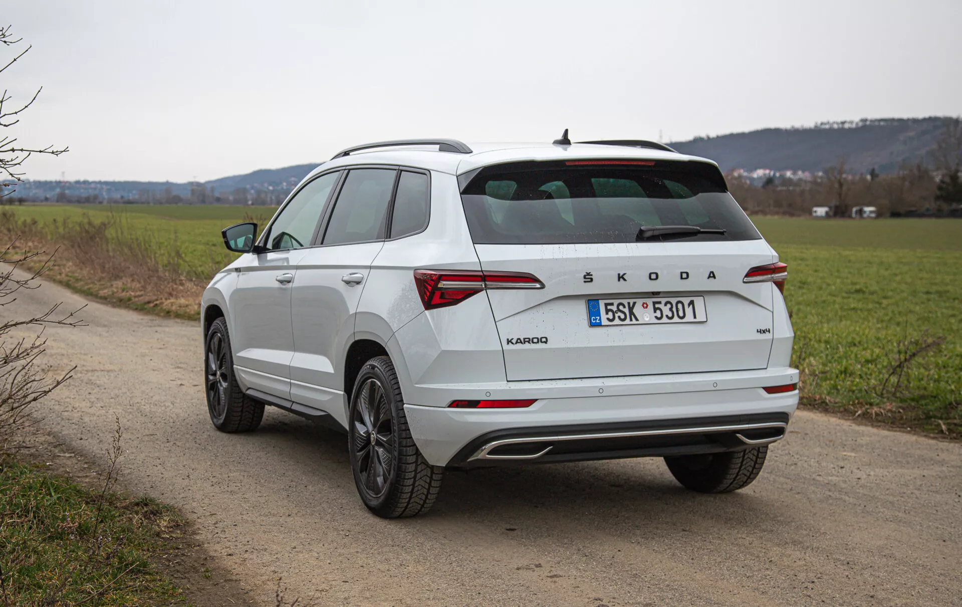 Vista posteriore a tre quarti di un SUV compatto Škoda Karoq bianco su una strada di campagna, con finestra posteriore inclinata, barre sul tetto, luci posteriori a LED, marchio Škoda, leghe a più razze e dettagli cromati, fotografato all'aperto durante il giorno con uno sfondo di paesaggio naturale.