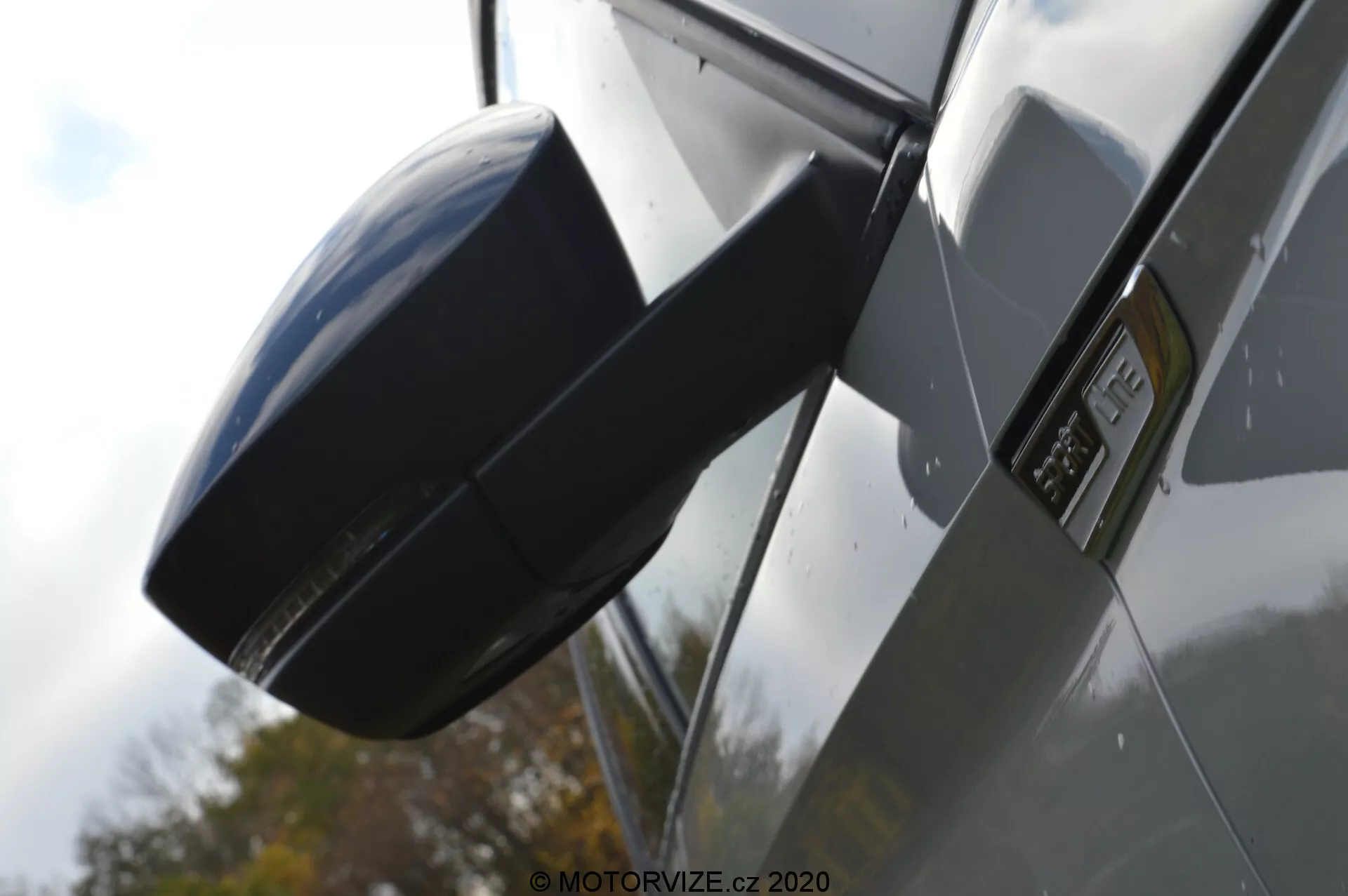 Vista laterale ravvicinata di una Skoda Kodiaq 2017 focalizzata sullo specchietto esterno e sulla zona del parafango. L'immagine è scattata da un'angolazione bassa, enfatizzando il profilo laterale della vettura, con una moderna calotta dello specchietto laterale aerodinamica in tinta con la carrozzeria del veicolo, un distintivo prominente sul parafango che indica un modello o livello di allestimento specifico, pannelli della carrozzeria lucidi e metallici e riflessi che suggeriscono un ambiente all'aperto con un'auto di colore scuro.