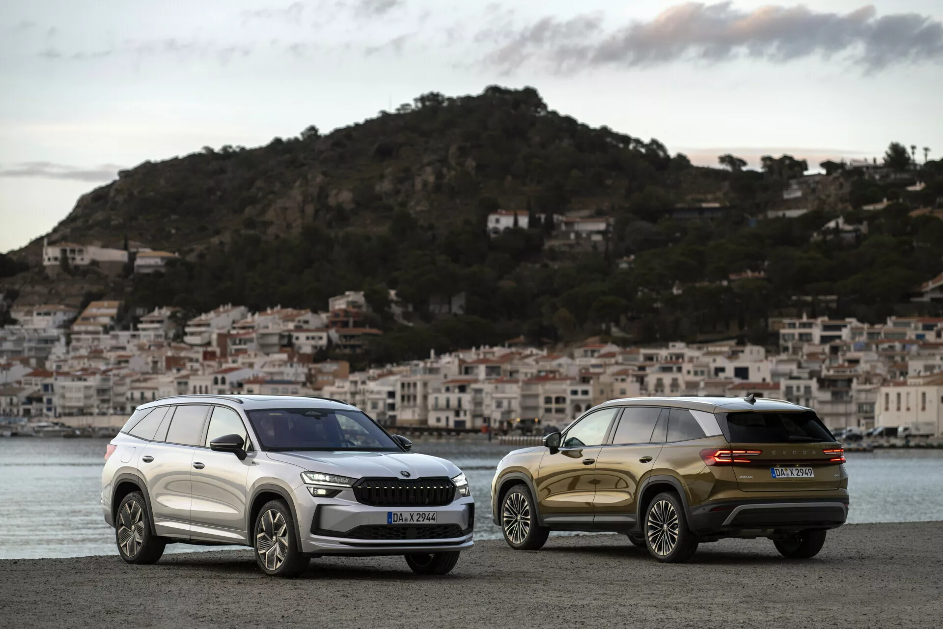 SUV Skoda Kodiaq 2024 parcheggiate una accanto all'altra all'alba su un lungomare con sfondo di un paese costiero, mostrando la griglia frontale, i fari e le linee di design eleganti di una e la vista posteriore di tre quarti dell'altra.