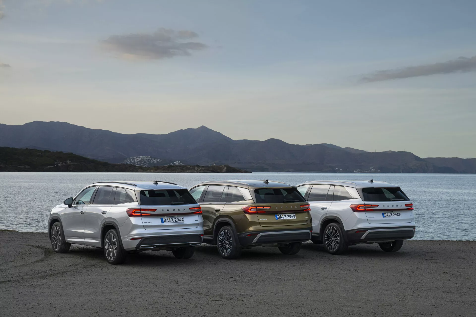 Lineup di Skoda Kodiaq 2024 con tre SUV parcheggiate sul lungomare durante il crepuscolo, mostrando i profili posteriori e l'estetica del design contro il sfondo di un paesaggio illuminato dolcemente