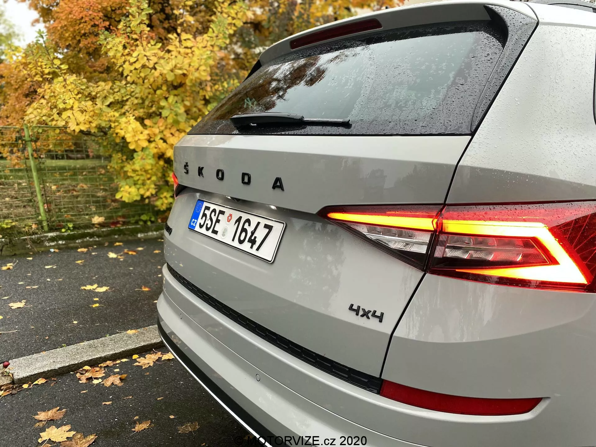 Vista posteriore di un veicolo Škoda argento con fanali posteriori a LED a forma di C illuminati, emblema e marchio Škoda, con badge 4x4, catturato dal lato posteriore destro su un marciapiede bagnato tra foglie autunnali e alberi.