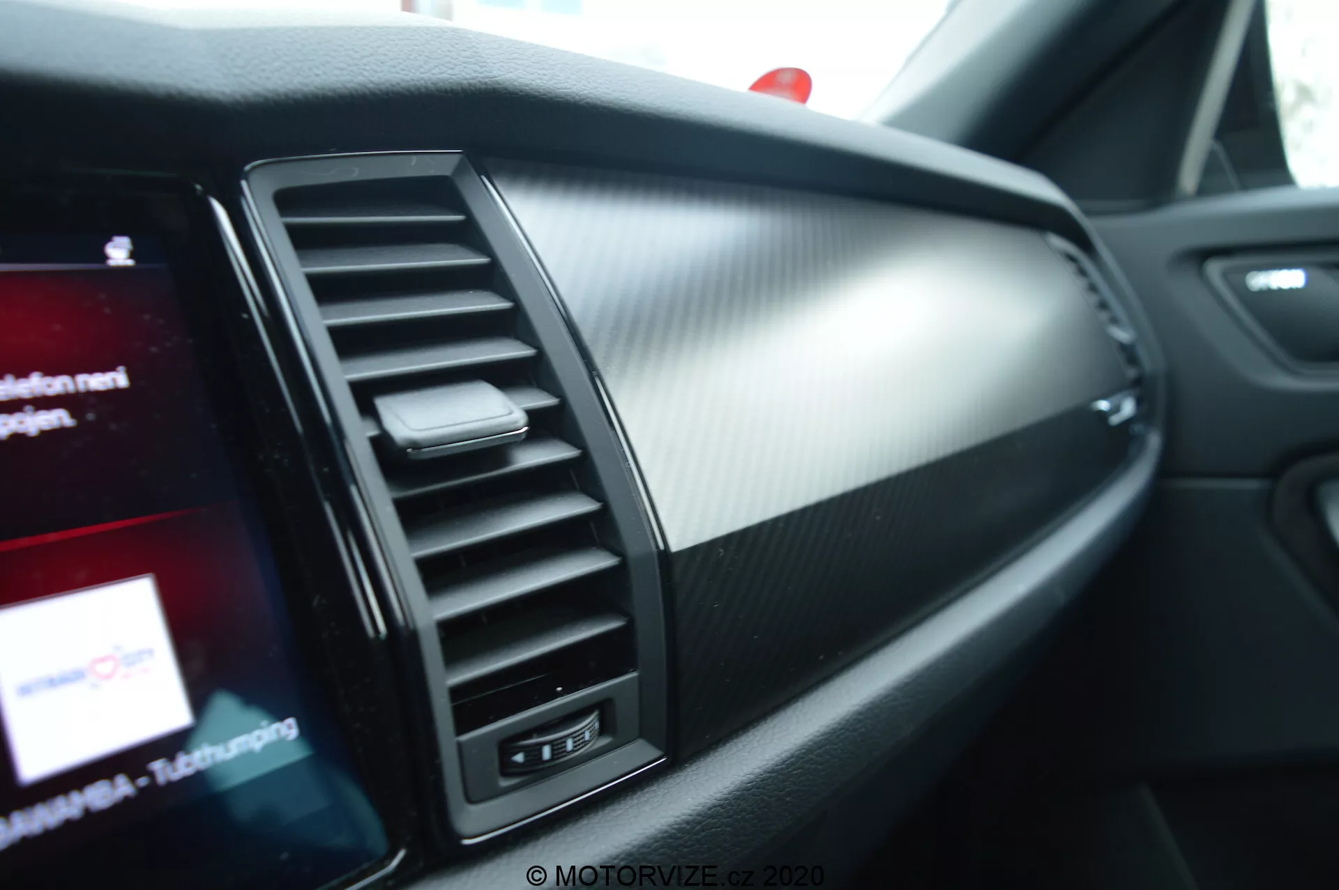 Vista ravvicinata dell'interno della Skoda Kodiaq 2017, evidenziando il lato passeggero del cruscotto. Caratteristiche includono una bocchetta di ventilazione con lamelle regolabili verticalmente, una sezione decorativa e uno schermo di infotainment parzialmente visibile con grafica colorata. I materiali del cruscotto combinano metallo spazzolato e plastica testurizzata che ricorda la fibra di carbonio, enfatizzando un design moderno e sportivo.