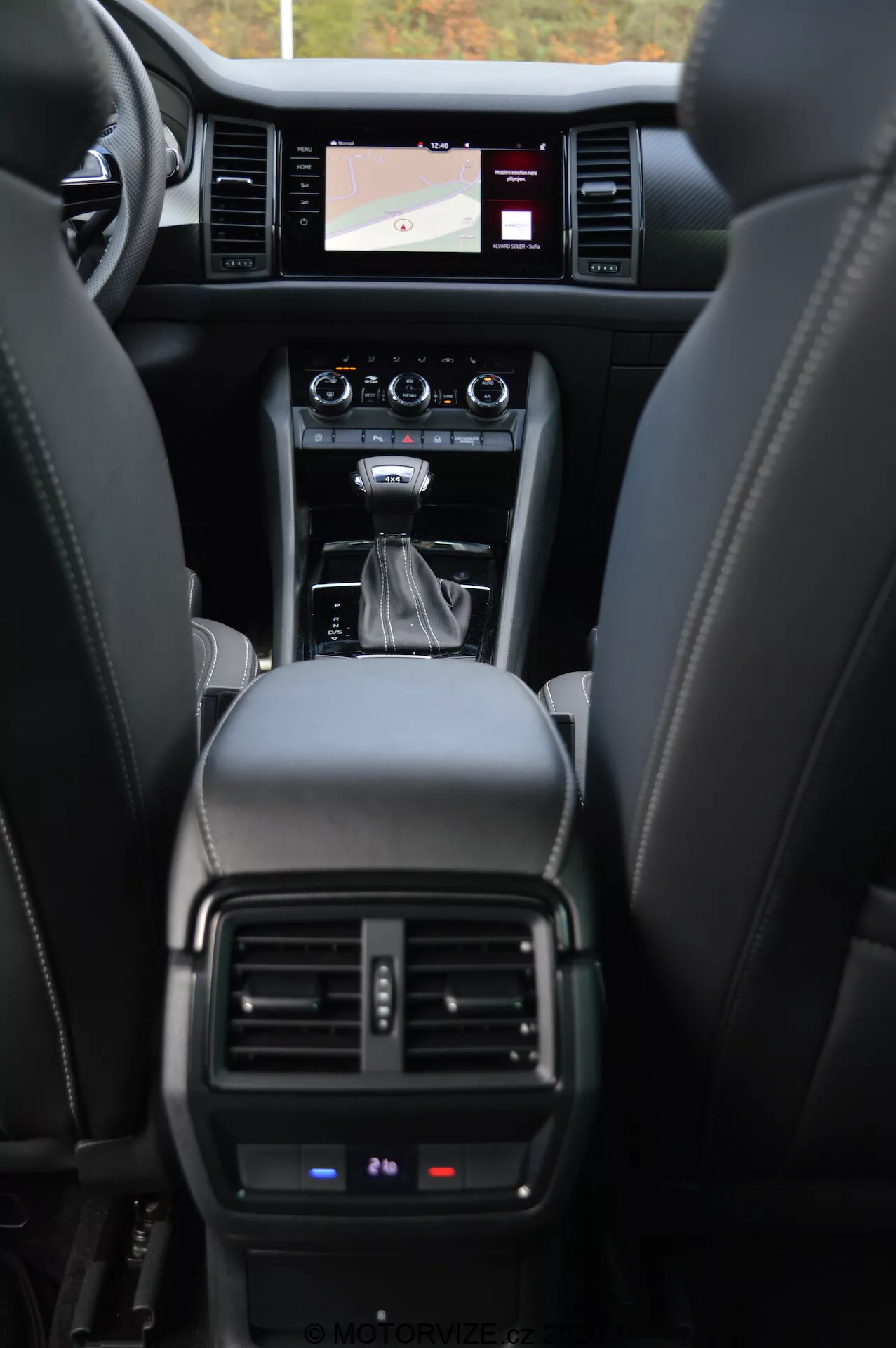 Interni della Skoda Kodiaq 2017, che mostra il cruscotto anteriore dalla prospettiva del sedile posteriore. L'immagine mette in evidenza i sedili in pelle, la console centrale con leva del cambio automatico, pulsanti di controllo, sistema di infotainment con schermo tattile che mostra una mappa di navigazione, bocchette di aerazione del climatizzatore e una vista parziale del quadro strumenti che mostra dettagli operativi critici.