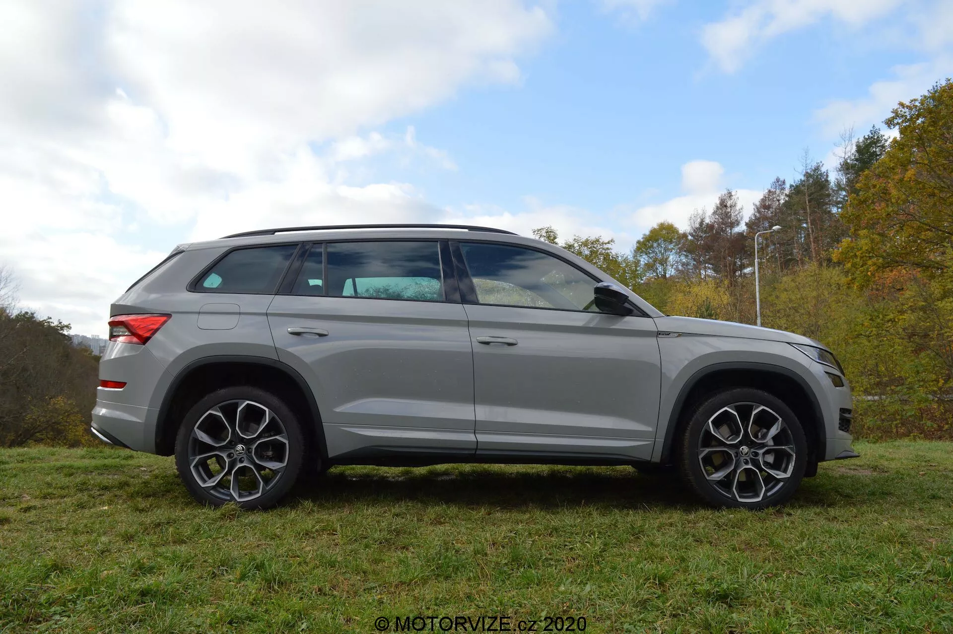Vista laterale completa di un SUV Skoda Kodiaq 2017 di colore argento parcheggiato sull'erba con uno sfondo di alberi e cielo nuvoloso. Il design del veicolo presenta linee audaci, grandi ruote in lega e distintivi cerchi multiraggi. Gli specchietti, che hanno indicatori di direzione integrati, sono montati appena sotto la linea dei finestrini. I vetri sono leggermente oscurati e la linea del tetto si inclina dolcemente verso la parte posteriore, indicando un profilo sportivo, simile a un coupé. Il SUV è anche dotato di un sistema di barre sul tetto.