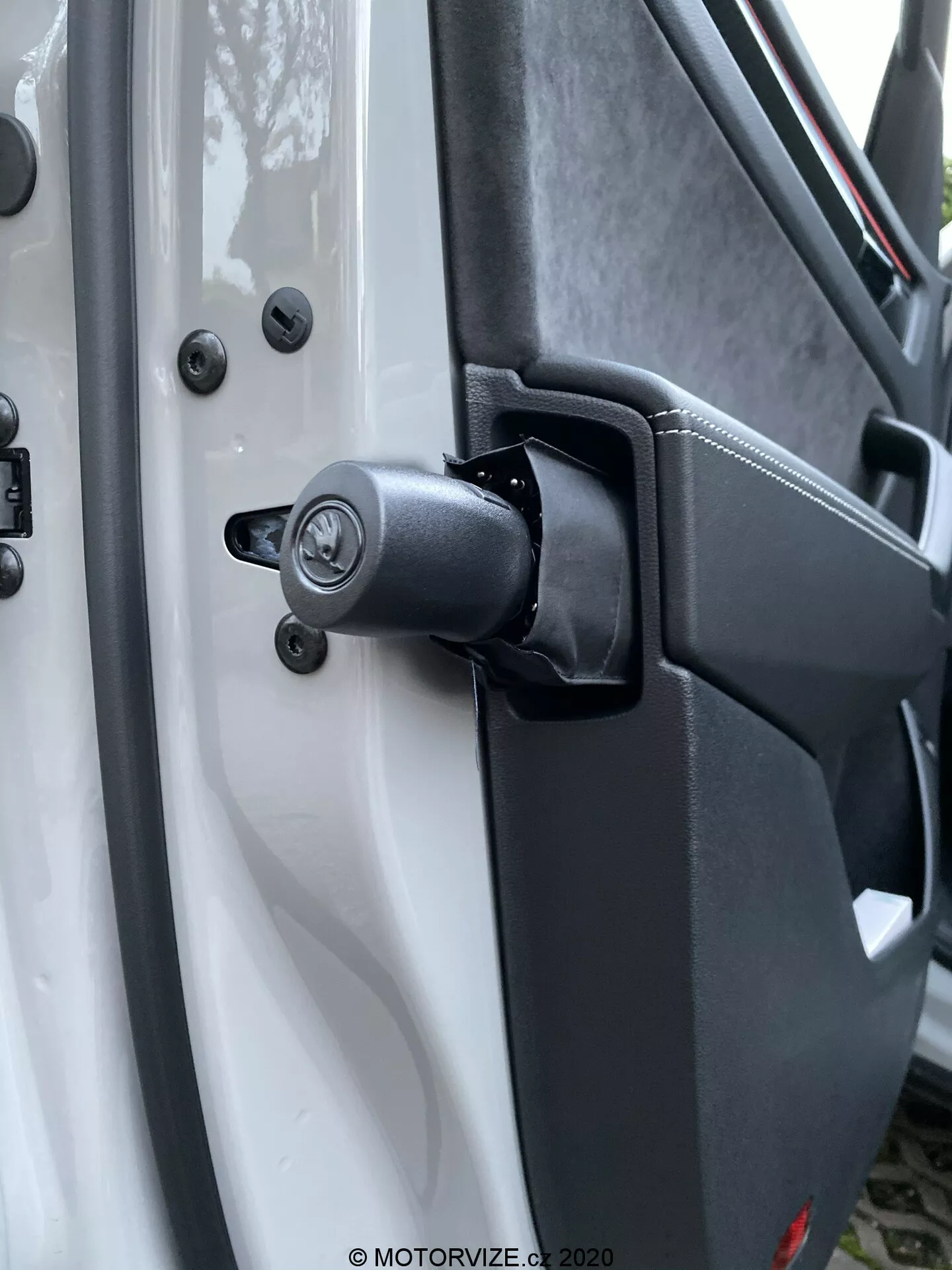 Vista ravvicinata del robusto meccanismo della cerniera della porta e dell'area circostante di una Škoda Kodiaq 2017, con una componente a molla visibile per uso intensivo. Il veicolo bianco contrasta con i componenti della cerniera neri, inclusi un meccanismo di chiusura tradizionale con serratura sopra la cerniera, fissato con bulloni a testa esagonale per la durata. La plastica testurizzata del pannello interno della porta e i punti di montaggio sicuri migliorano il design utilitario. Gli elementi di sfondo sfocati evidenziano la cerniera e il meccanismo di chiusura.