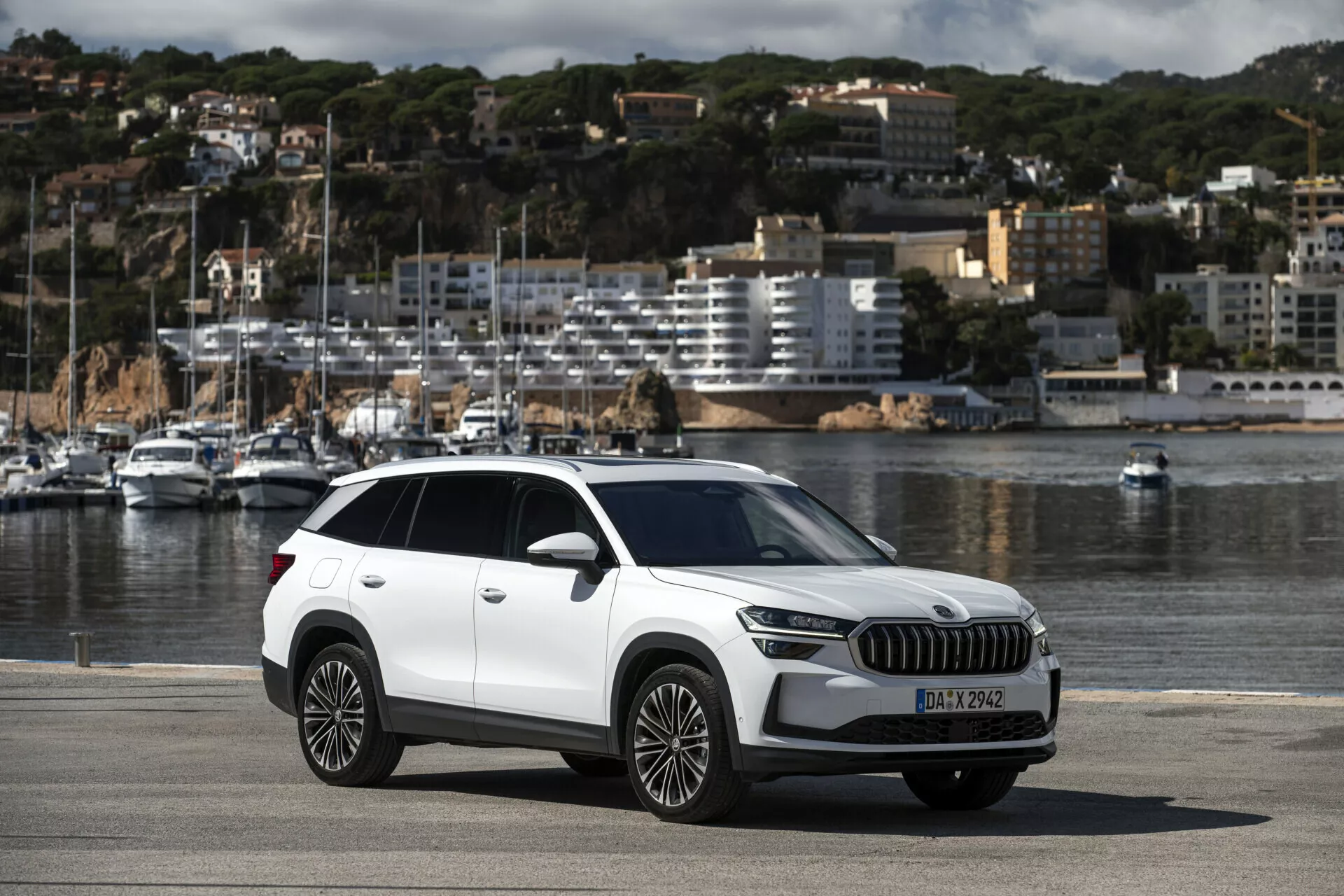SUV crossover Skoda Kodiaq 2024 parcheggiata su un lungomare che mostra la vista completa dell'auto inclusi elementi di design moderno come fari LED, grande griglia, ruote in lega a più razze, con scenico sfondo di barche e paesaggio urbano