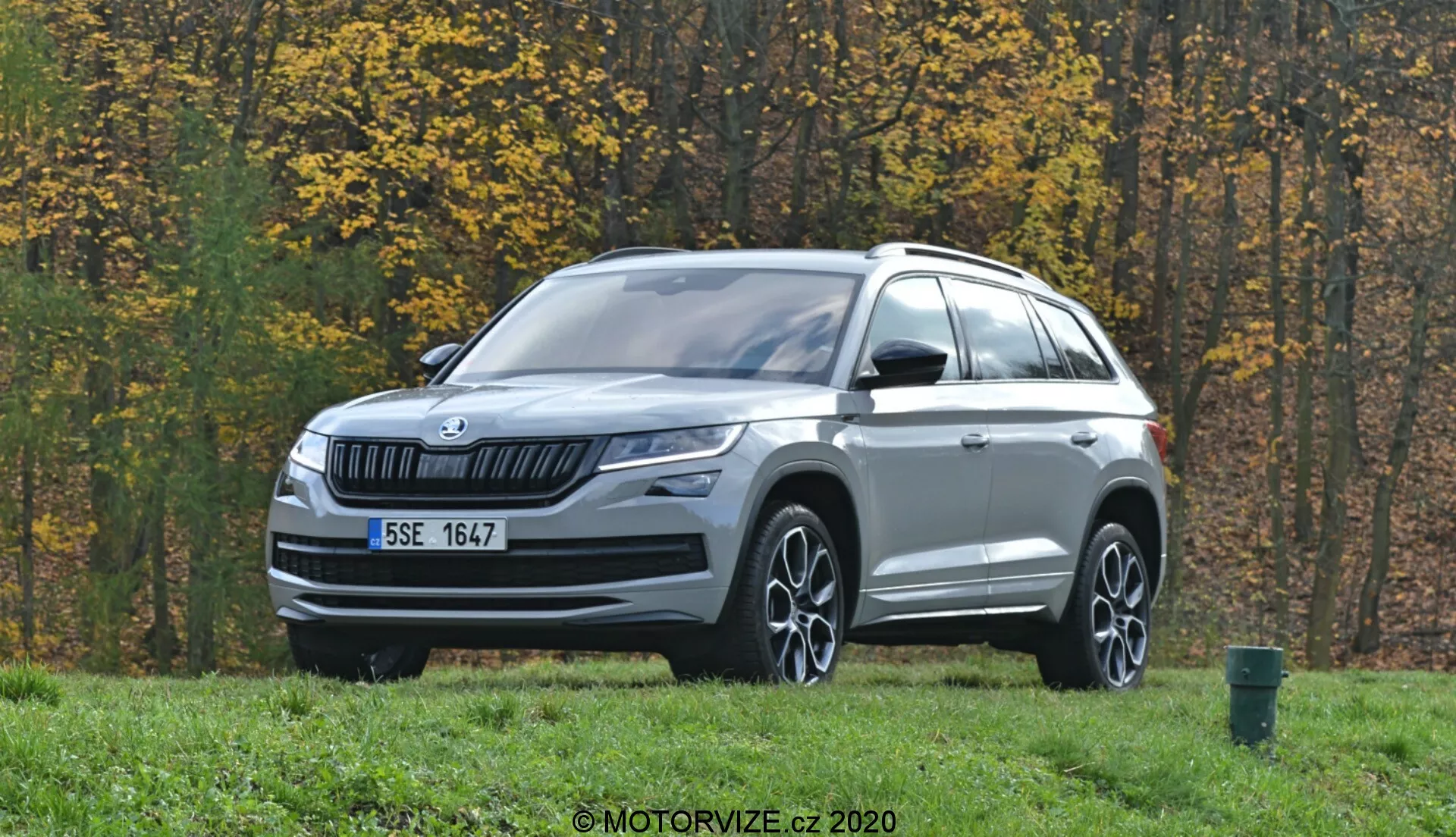 L'immagine mostra un SUV Skoda Kodiaq 2017 parcheggiato su un'area erbosa con alberi autunnali sullo sfondo. Fotografato da un'inquadratura frontale a tre quarti, il veicolo presenta una griglia frontale con lamelle orizzontali con il logo Skoda al centro. I fari LED eleganti si estendono verso i lati, accompagnati da fari fendinebbia sul paraurti inferiore. Il cofano ha linee sottili e una finitura argentata decora la parte inferiore del paraurti anteriore. Cerchi in lega con design multirazza, specchietti laterali con indicatori di direzione integrati, barre sul tetto e linee scolpite nel corpo evidenziano il profilo. I sensori o le telecamere nella parte inferiore del paraurti suggeriscono sistemi avanzati di assistenza alla guida (ADAS). L'ambiente esterno include una strada asfaltata e un cestino dei rifiuti verde sul bordo destro dell'immagine.