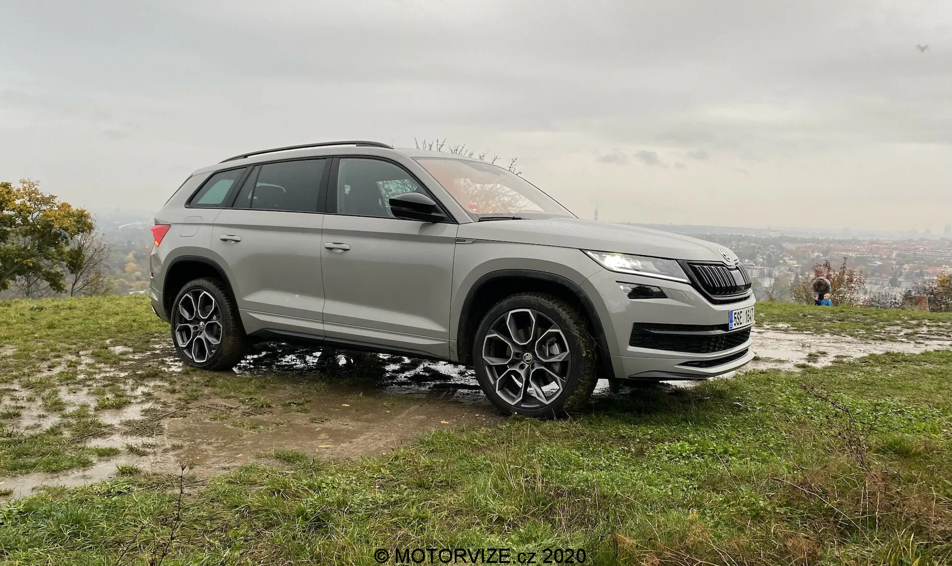Scatto laterale di un SUV moderno preso da un angolo obliquo che mostra sia gli elementi di design anteriori che laterali, ambientato in uno sfondo naturale da un'altura elevata. L'immagine mette in risalto la griglia anteriore, i fari, i fendinebbia e il logo distintivo del veicolo. Presenta anche un'elevata altezza da terra, grandi ruote in lega con design multispiro, arcate delle ruote prominenti, linea di spalla alta, specchietti laterali, barre del tetto e vetri oscurati. Il design sportivo e le potenziali tecnologie avanzate del veicolo sono accentuati in questo scatto stazionario diurno con macchie di neve, indicando una stagione più fredda.