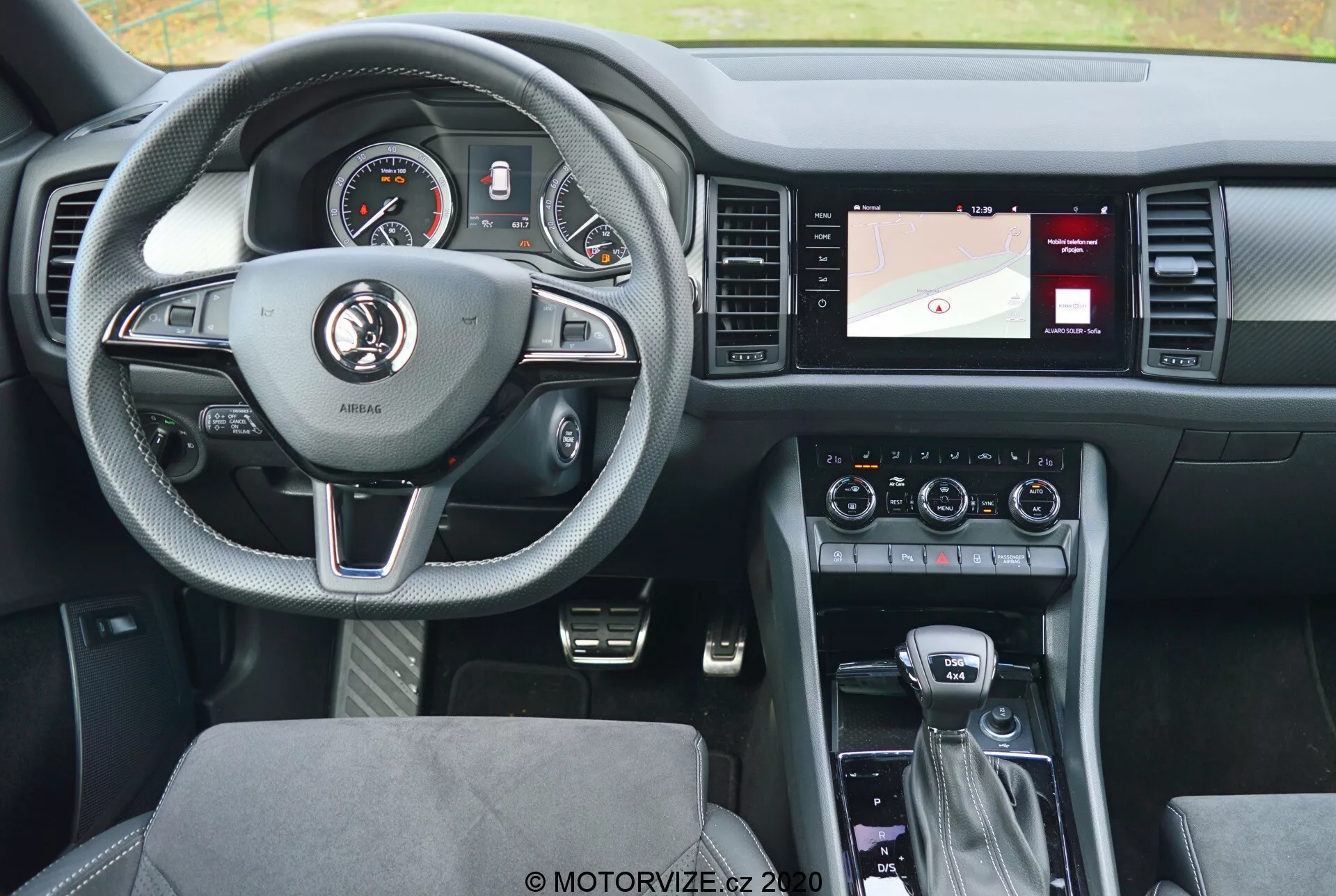 Vista interna di una Skoda Kodiaq 2017 dal sedile posteriore, che evidenzia il volante a tre razze rivestito in pelle con dettagli argentati e pulsanti di controllo per il cruise control, l'audio e la comunicazione a mani libere. Il cruscotto dietro il volante presenta quadranti analogici e un display digitale. La console centrale a destra include uno schermo con una mappa per la navigazione, controlli per il clima e le impostazioni audio. La leva del cambio indica una trasmissione automatica, con ulteriori controlli per il sistema di infotainment e le modalità di guida. Il design moderno mira a un equilibrio tra comfort e tecnologia, con sedili rivestiti in tessuto a motivi e una disposizione ben illuminata e user-friendly.