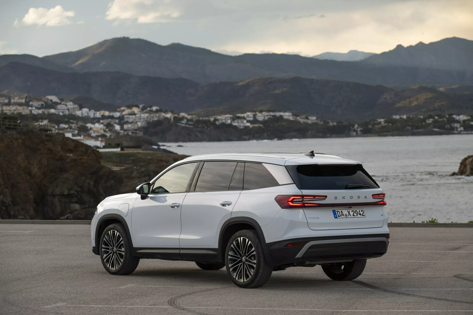 Vista posteriore a tre quarti di una Skoda Kodiaq 2024 di colore bianco, che mostra le linee di design laterali e posteriori, cerchi in lega a più razze, fanali posteriori LED e il badge del modello, parcheggiata all'aperto su una strada costiera all'ora dorata