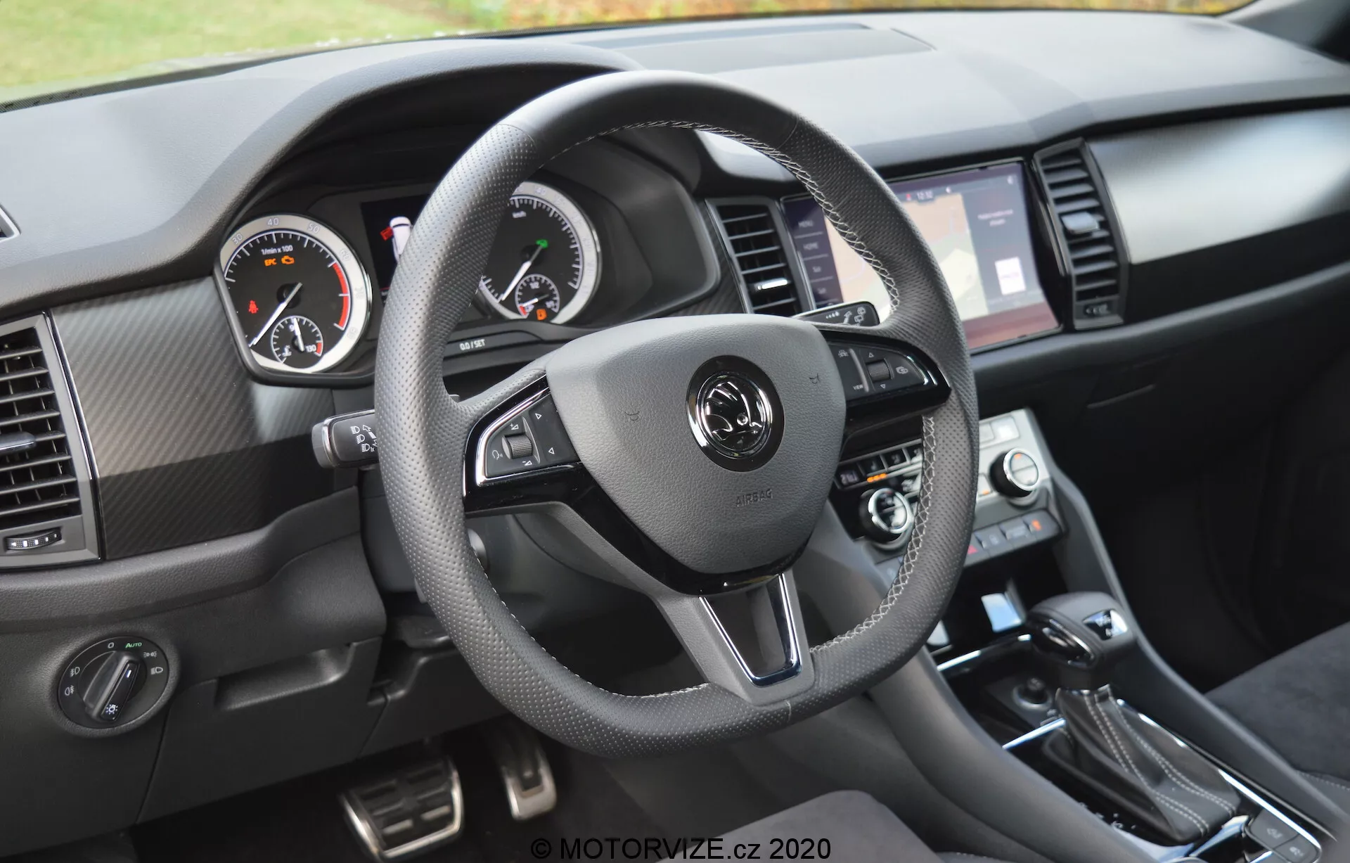 Vista interna di una Skoda Kodiaq 2017 focalizzata sulla prospettiva del conducente del cruscotto. Il volante a tre razze, rivestito in materiale simile a pelle con cuciture a contrasto, presenta pulsanti integrati per il controllo audio, la comunicazione a mani libere e le impostazioni del cruise control. Gli indicatori analogici, con il tachimetro a sinistra e il contagiri a destra, sono affiancati da un display digitale al centro. Il touchscreen di infotainment, affiancato da prese d'aria, manopole di controllo del clima e leva del cambio, sono visibili, insieme a materiali testurizzati e accenti metallici spazzolati.