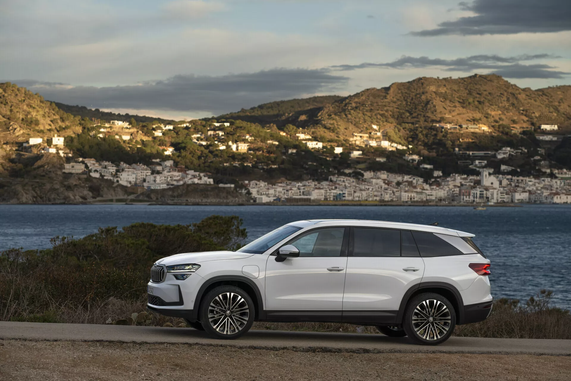 SUV moderno Skoda Kodiaq 2024 parcheggiata su una strada costiera con vista panoramica su una baia e case sul lato della collina, caratterizzata da un design elegante, passo lungo, elevata altezza da terra, linee della carrozzeria nitide, maniglie delle porte a scomparsa, vetri posteriori oscurati, barre sul tetto, grandi ruote in lega, fari LED, specchietti laterali con indicatori di direzione e tetto panoramico, tutto su una finitura esterna bianca a contrasto.