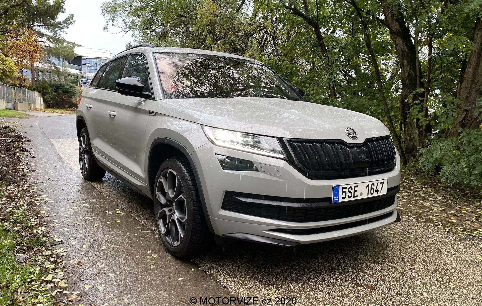 Skoda Kodiaq 2017 SUV moderno di un marchio automobilistico europeo, angolazione frontale-complessiva che mostra il profilo anteriore e laterale fino a metà della portiera posteriore, parcheggiato lungo un sentiero fiancheggiato da erba e alberi in un ambiente urbano o suburbano. Le caratteristiche includono una griglia anteriore caratteristica con lamelle verticali, fari LED eleganti, fari fendinebbia nel paraurti, grandi prese d'aria sul paraurti anteriore, linee di contorno sul cofano per l'aerodinamica, passaruota prominenti con cerchi in lega a più raggi, specchietti laterali con indicatrici di direzione integrate, modanature protettive sul lato inferiore e verniciatura nel complesso pulita e ben mantenuta.