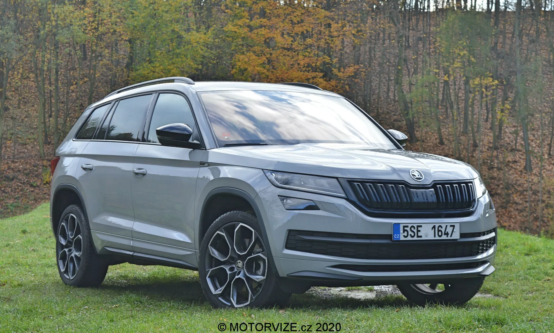 Questa immagine mostra un SUV Škoda Kodiaq del 2017 catturato da un angolo frontale di tre quarti, focalizzandosi sul lato sinistro del veicolo. Lo scatto mette in evidenza la griglia anteriore con il caratteristico design Škoda a lamelle verticali, la configurazione dei fari probabilmente con tecnologia LED e il logo del marchio al centro. Il profilo laterale include la portiera anteriore del conducente, la portiera posteriore del passeggero, le barre sul tetto e parti del quarto posteriore. Sono ben visibili anche i cerchi in lega caratterizzati da un design a raggi. L'ambientazione esterna con sfondo erboso e fogliame autunnale esalta l'estetica del moderno SUV.