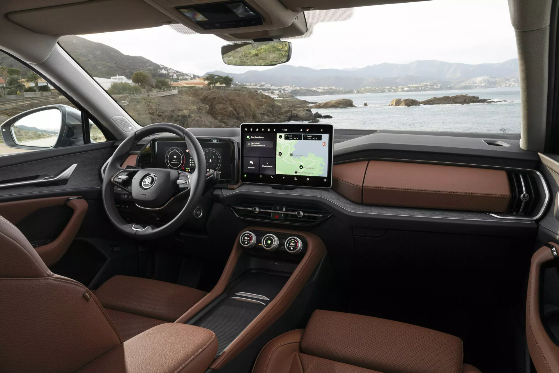 Vista interna della Skoda Kodiaq 2024 dalla prospettiva del passeggero che mostra il cruscotto, il volante multifunzione con logo, il quadro strumenti digitale, il grande sistema di infotainment touchscreen con GPS, sedili lussuosi in pelle marrone e il design raffinato della consolle centrale, impostato contro un tranquillo paesaggio costiero fuori dalle finestre