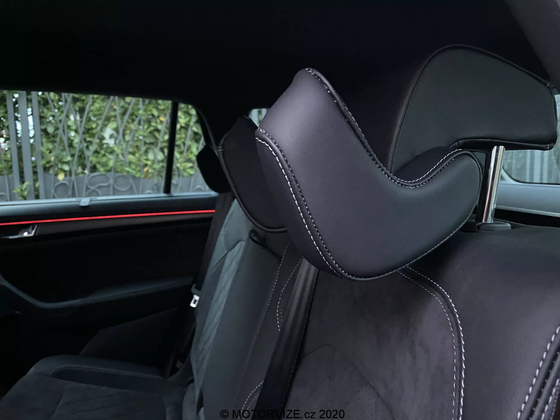 Vista interna di una Skoda Kodiaq 2017 focalizzata su un sedile dell'auto. La ripresa angolata enfatizza la porzione del poggiatesta, rivestita in materiale nero, probabilmente pelle o sintetico di alta qualità, con un contorno distinto per il comfort. Le cuciture precise con filo a contrasto aggiungono accenti lussuosi. Lo sfondo rivela una luce ambientale rossa orizzontale, che mette in risalto gli elementi del design automobilistico moderno. I servizi visibili per i passeggeri posteriori includono cinture di sicurezza e forse un bracciolo centrale, indicando la seconda fila di sedili.