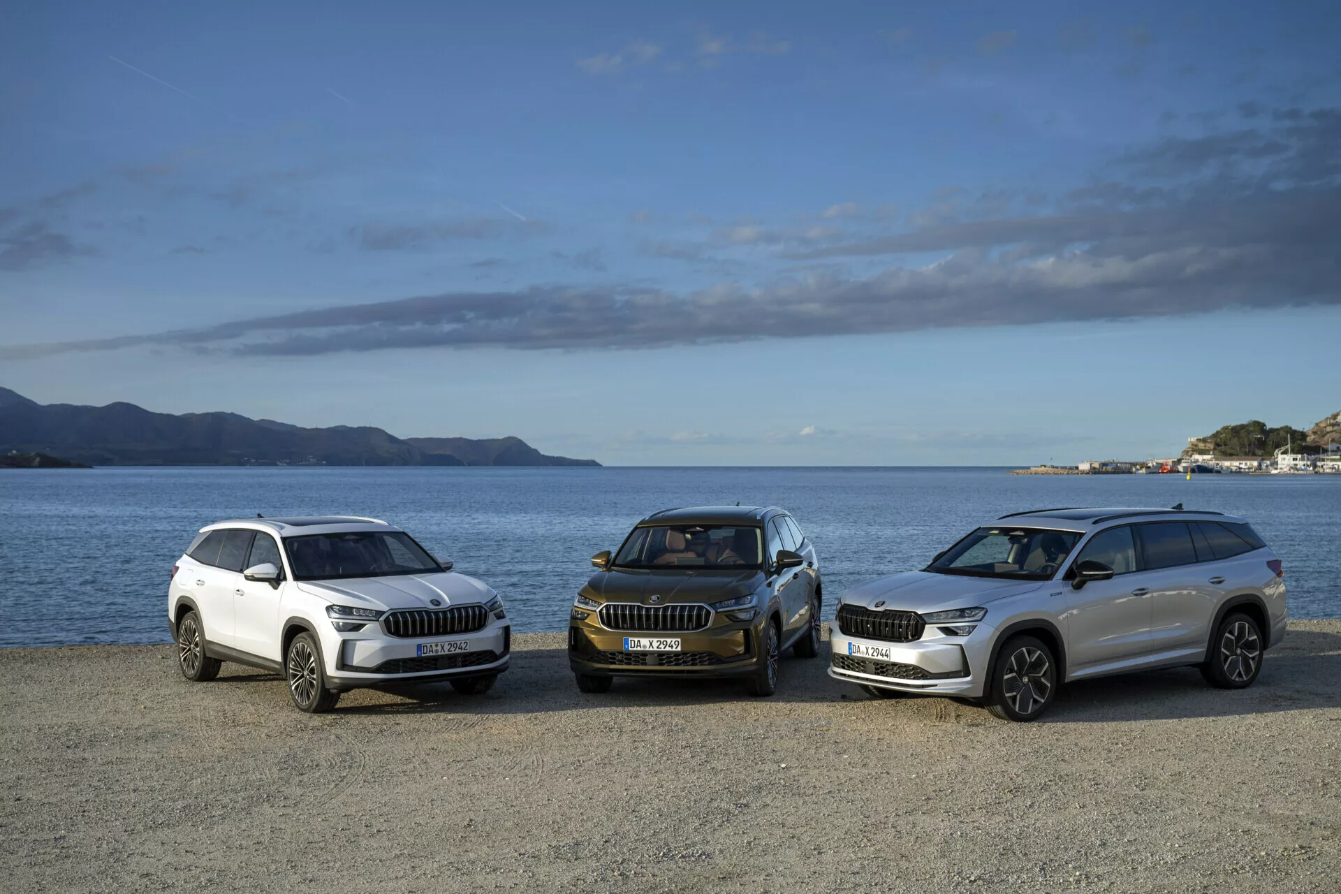 Gamma di SUV Skoda Kodiaq 2024 in colori bianco e scuro in vista frontale laterale, parcheggiate su un lungomare con montagne e un cielo parzialmente nuvoloso sullo sfondo