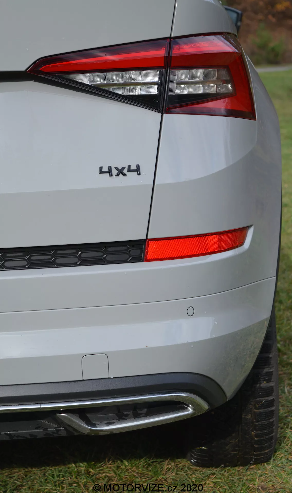 Vista ravvicinata del quarto posteriore sinistro di una Skoda Kodiaq 2017. L'immagine si concentra sul fanale posteriore, con luci LED moderne dal design angolare e nitido e chiare suddivisioni per diverse funzioni di luce. Il badge '4X4' indica la trazione integrale. Il paraurti posteriore include riflettori e sensori, probabilmente per l'assistenza al parcheggio, e un terminale di scarico visibile. La vernice argento metallica e le linee sottili della carrozzeria sono visibili, con uno sfondo di erba e alberi che suggerisce un'ambientazione suburbana all'aperto. Filigrana 'MOTORVIZE.cz 2020'.