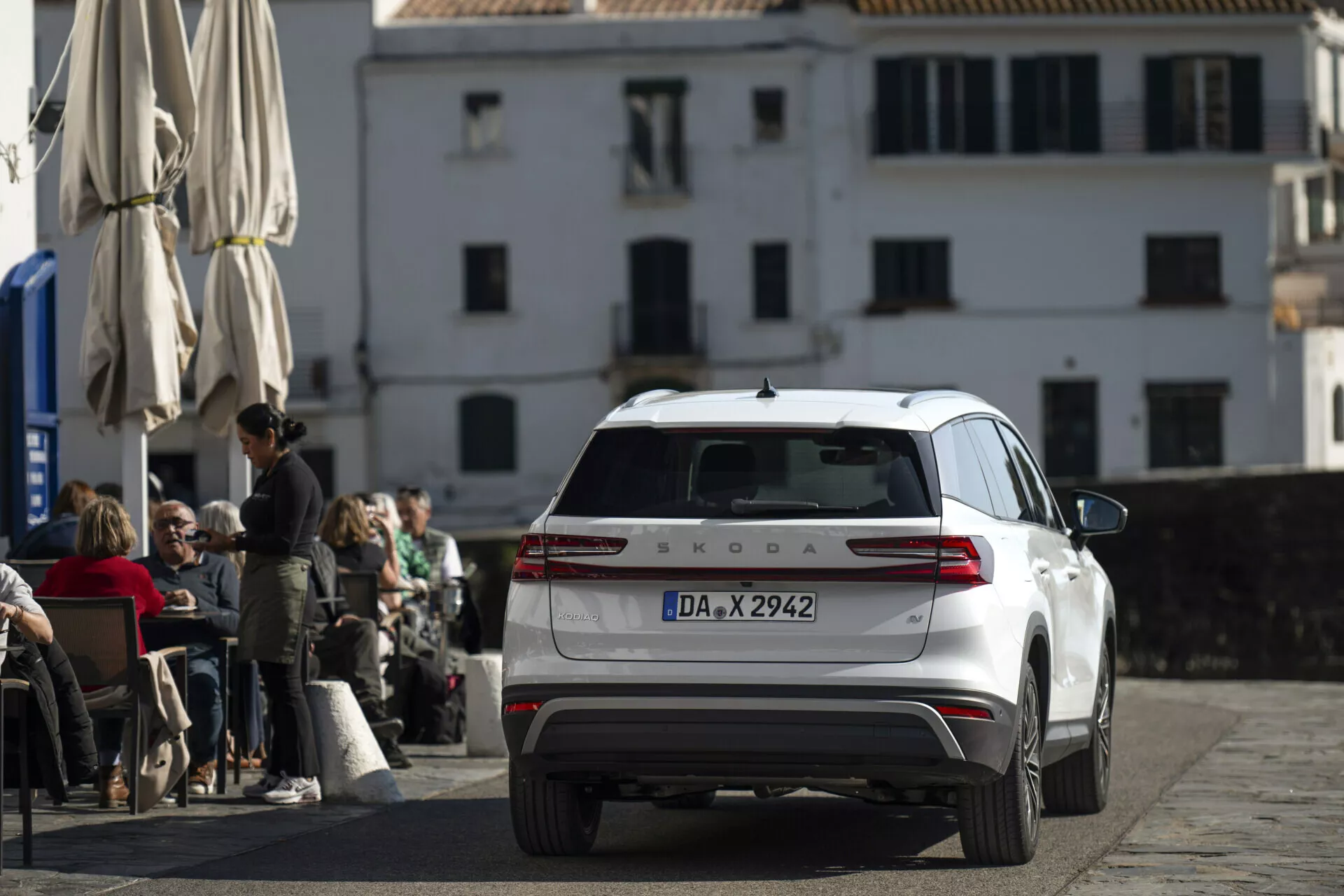 Škoda Kodiaq bianca 2024 fotografata da un angolo posteriore e leggermente rialzato, mostrando la parte posteriore completa, luci posteriori, sportello del bagagliaio, finestra posteriore con tergicristallo e tetto con antenna e barre, parcheggiata su una strada acciottolata di fronte a un caffè con architettura europea classica sullo sfondo.